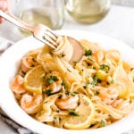 A bowl of Easy Shrimp Scampi with Linguine is topped with lemon slices and fresh herbs, while a hand twirls pasta on a fork. Two glasses of white wine are in the background, completing this inviting scene.