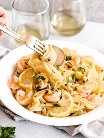 A bowl of Easy Shrimp Scampi with Linguine is topped with lemon slices and fresh herbs, while a hand twirls pasta on a fork. Two glasses of white wine are in the background, completing this inviting scene.