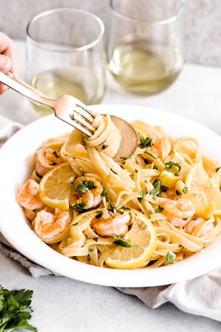 A bowl of Easy Shrimp Scampi with Linguine is topped with lemon slices and fresh herbs, while a hand twirls pasta on a fork. Two glasses of white wine are in the background, completing this inviting scene.
