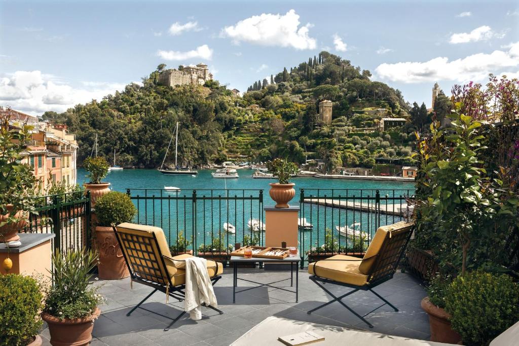 A terrace with two cushioned chairs and a table overlooks a harbor with boats, clear turquoise water, and lush green hills with villas—an inviting scene reminiscent of Portofino boutique hotels under a partly cloudy sky.