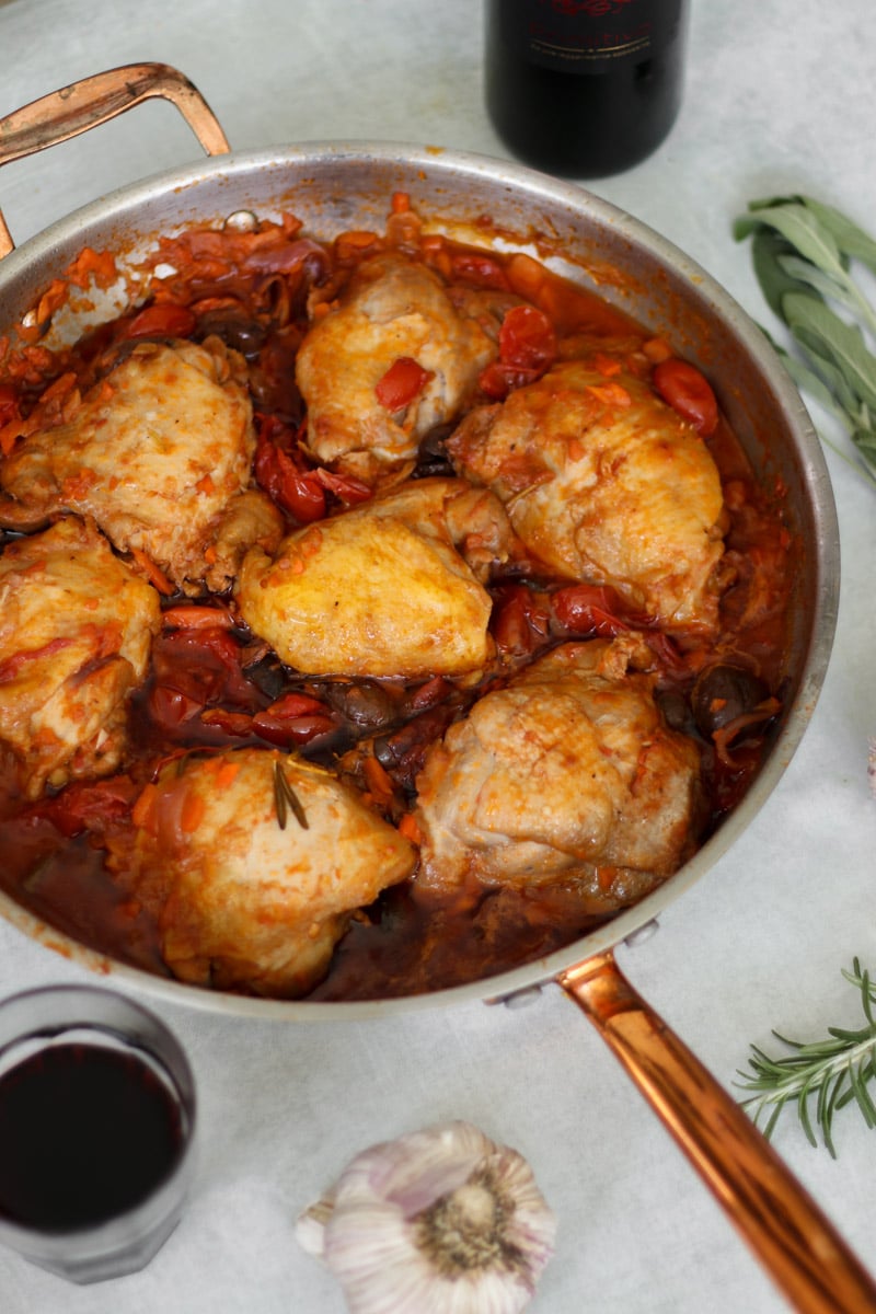 overhead image of chicken cacciatore in a skillet with fresh herbs and wine in background.