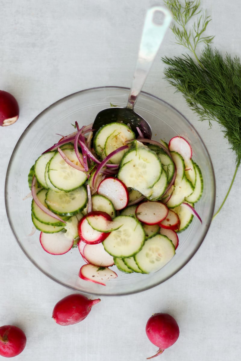 overhead image of Mediterranean cucumber salad in a glass bowl with herbs and radishes in background.