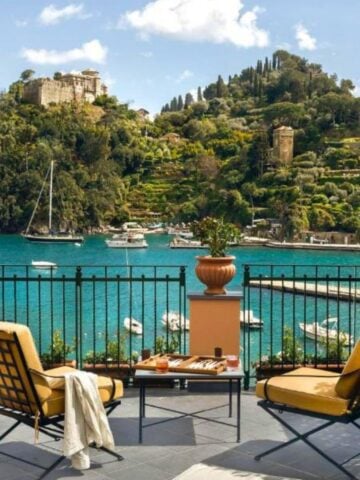 A seaside terrace with two yellow cushioned chairs and a table overlooks a turquoise bay dotted with boats, lush green hills, villas, and a historic stone building—an idyllic scene reminiscent of Portofino boutique hotels.