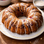 A round strawberry bundt cake with visible pieces of fruit, topped with white icing drizzle, sits on a white plate on a wooden table. Fresh strawberries and a bowl of glaze are in the background.