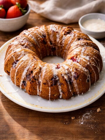 A round strawberry bundt cake with visible pieces of fruit, topped with white icing drizzle, sits on a white plate on a wooden table. Fresh strawberries and a bowl of glaze are in the background.
