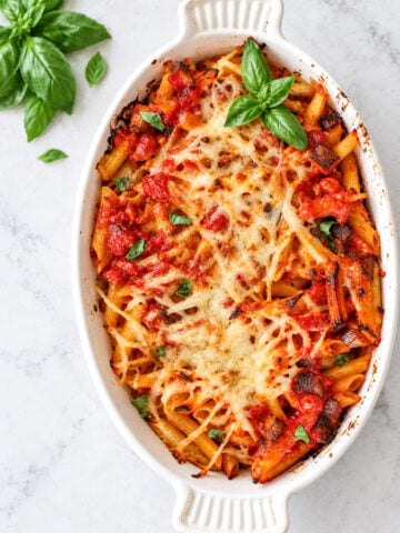 Baked Pasta with Tomatoes and Eggplant in a white oval dish topped with melted cheese, tomato sauce, and fresh basil, with basil sprigs on the side.