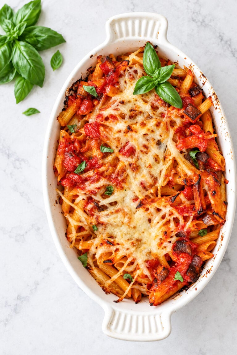 Baked Pasta with Tomatoes and Eggplant in a white oval dish topped with melted cheese, tomato sauce, and fresh basil, with basil sprigs on the side.