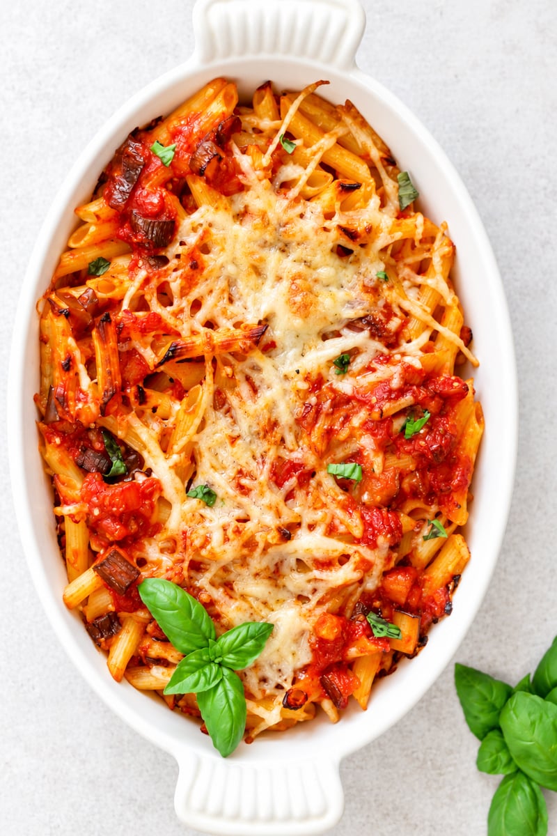Baked Pasta with Tomatoes and Eggplant topped with melted cheese and tomato sauce, garnished with fresh basil leaves, served in a white oval dish on a light background.
