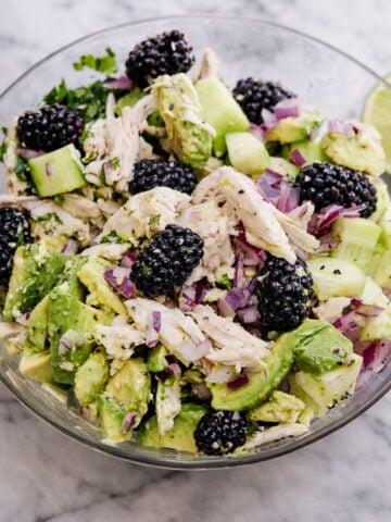 A glass bowl filled with Chicken Avocado Salad, featuring shredded chicken, chopped avocado, cucumber, red onion, fresh cilantro, and whole blackberries on a marble surface.
