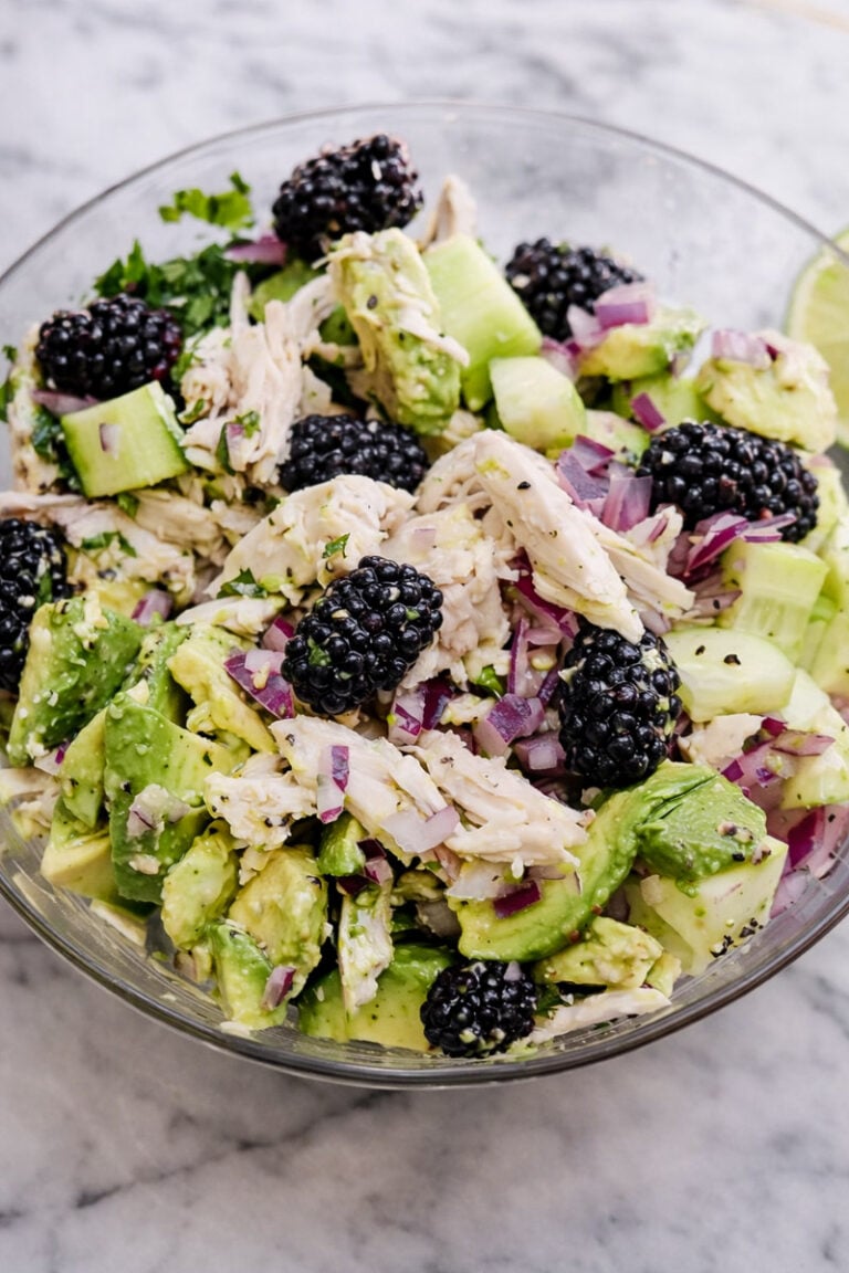 A glass bowl filled with Chicken Avocado Salad, featuring shredded chicken, chopped avocado, cucumber, red onion, fresh cilantro, and whole blackberries on a marble surface.