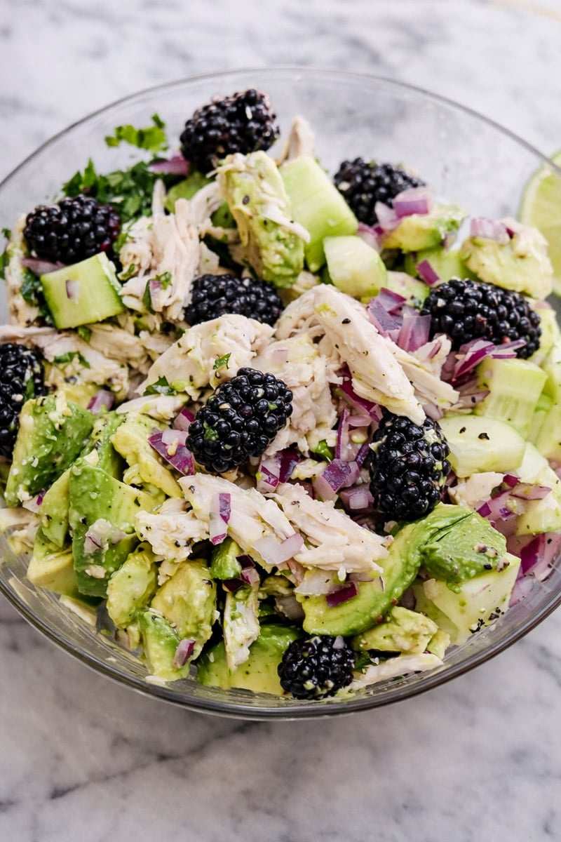 A glass bowl filled with Chicken Avocado Salad-shredded chicken, avocado chunks, cucumber, diced red onion, fresh blackberries, and herbs-all arranged on a marble surface.
