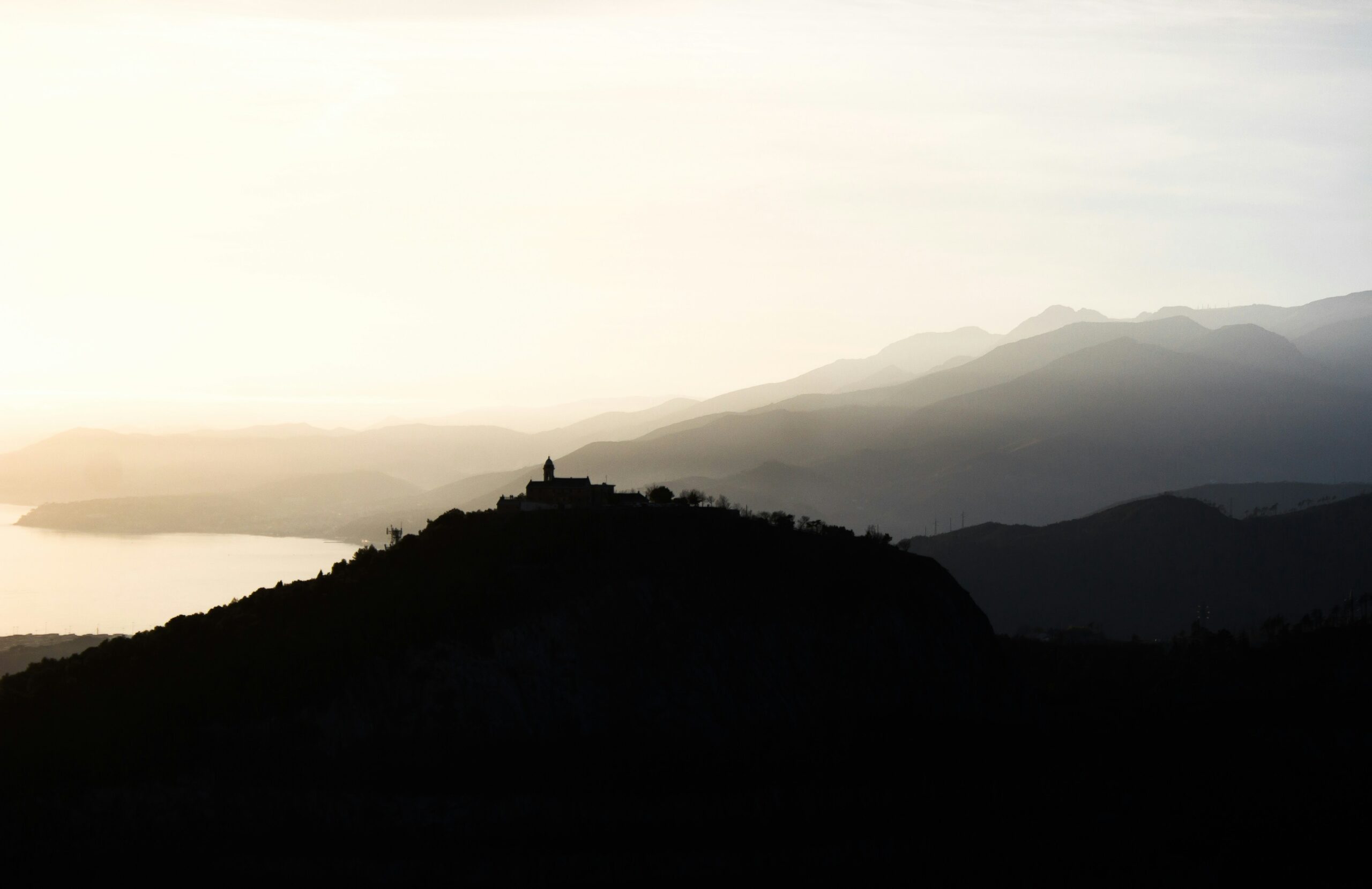 The photograph offers a breathtaking view of Monte Gazzo and the surrounding mountain skyline in the Riviera Ligure, Italy. The scene is dominated by the rugged, green-covered peaks of Monte Gazzo, which rises majestically against a clear blue sky. The mountains are part of the Ligurian Apennines, and their slopes are dotted with patches of dense forest and terraced hillsides typical of the region. Below, the landscape gently descends toward the coastline, with glimpses of quaint villages and the shimmering Ligurian Sea in the distance. The overall view is serene and picturesque, capturing the natural beauty of the Ligurian Riviera and its harmonious blend of mountains and sea.
