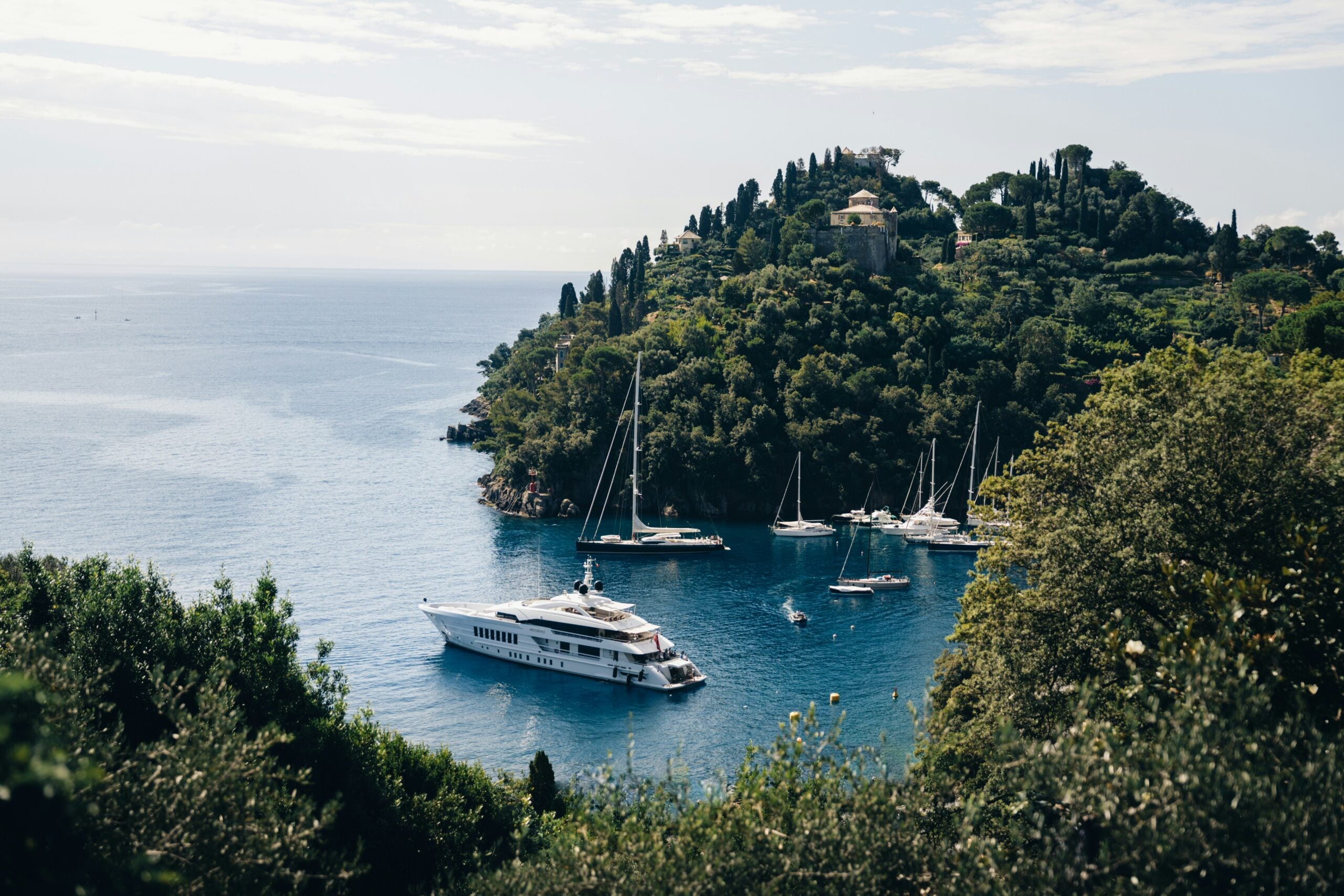 The photograph showcases the picturesque harbor of Portofino, a renowned village in the Metropolitan City of Genoa, Italy. The scene captures the vibrant, colorful buildings that line the waterfront, their facades painted in warm pastel shades that contrast beautifully with the deep blue waters of the Ligurian Sea. Elegant yachts and small fishing boats are anchored in the harbor, gently swaying in the calm waters. The lush green hills in the background add a natural frame to the scene, enhancing the idyllic charm of this Mediterranean gem. The overall atmosphere is one of serene luxury and timeless beauty, making Portofino a must-visit destination for travelers exploring the Genoa region.