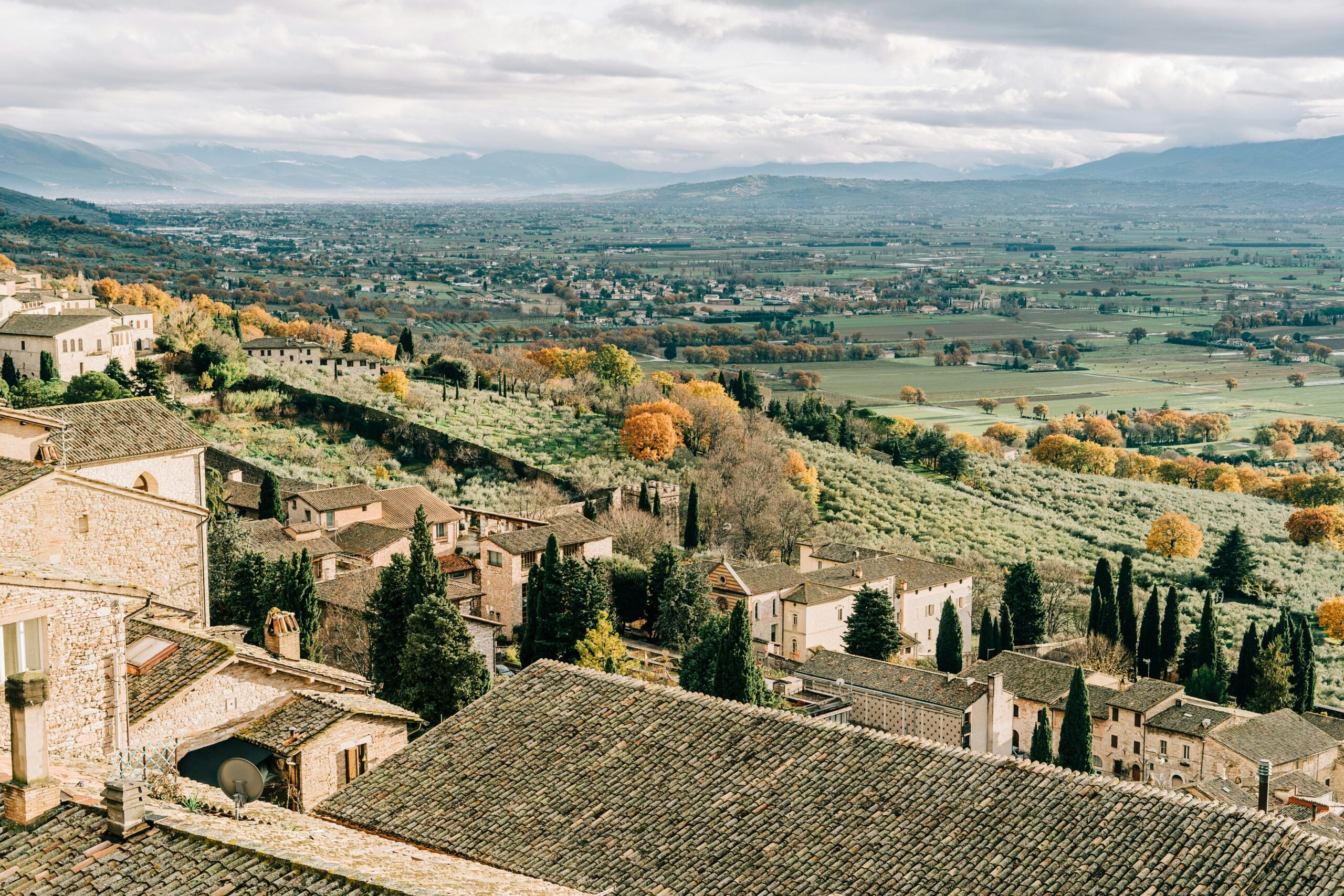 A beautiful village overlooking the countryside near Perugia, Italy, featuring charming stone houses with terracotta roofs and narrow winding streets. The village is perched on a hill, offering panoramic views of the lush green fields, vineyards, and rolling hills below. The serene landscape, bathed in golden sunlight, perfectly captures the idyllic and picturesque essence of the Umbrian countryside near Perugia.
