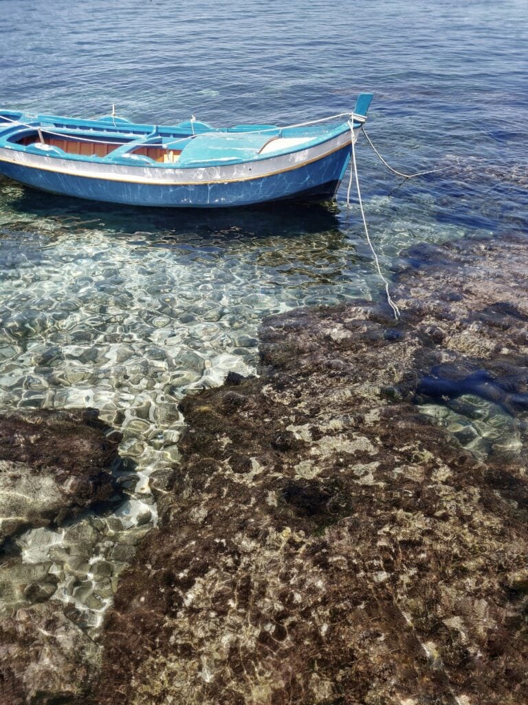 A photograph captures a small fishing boat docked in the clear blue, rocky waters of Palermo, Sicily. The scene highlights the serene coastal beauty and maritime traditions of Palermo, with the boat gently swaying against a backdrop of rugged rocks and azure waters. Keywords: Palermo, Sicily, small fishing boat, blue rocky waters.