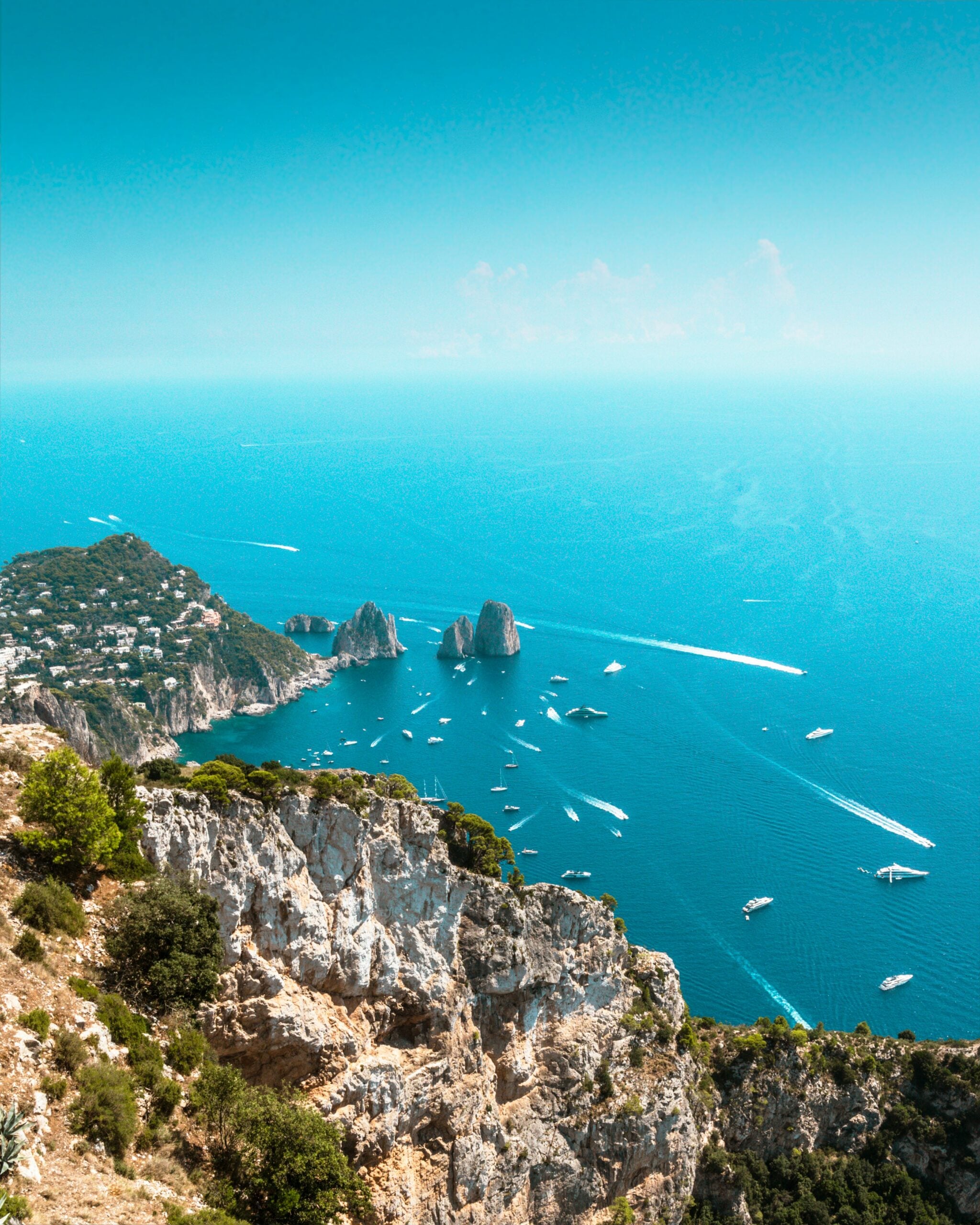 A stunning view from high atop the cliffs in Capri, Italy, overlooking boats gently bobbing near the shore. The picturesque scene captures the island's rugged cliffs descending into the crystal-clear blue waters of the Mediterranean Sea, where a variety of boats are anchored. This breathtaking vantage point on Capri Island, Italy, highlights the island's natural beauty and its appeal as a premier coastal destination.