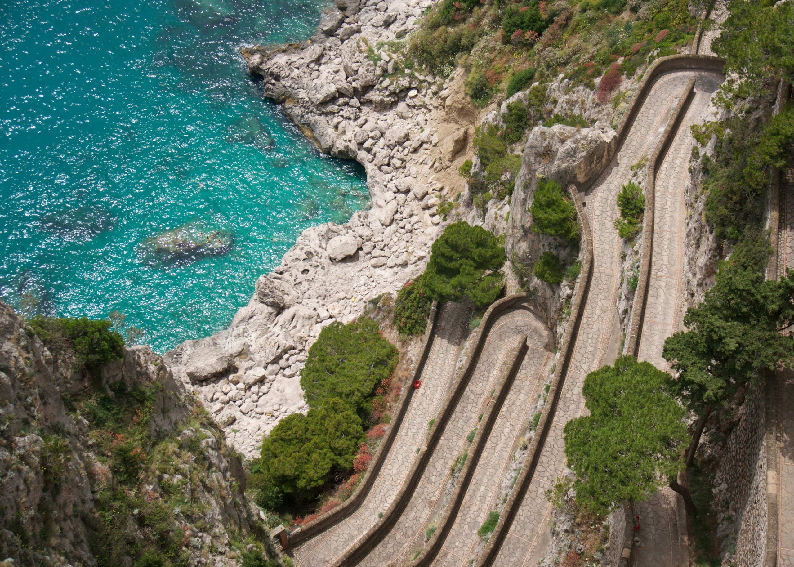 A breathtaking view of the steep serpentine road on Capri Island, Italy, captured from above. The winding road meanders through lush greenery and rugged cliffs, offering stunning vistas of the vibrant blue waters of the Tyrrhenian Sea. This iconic scene on Capri Island, Italy, highlights the island's dramatic landscape and natural beauty, making it a must-visit destination for travelers seeking picturesque coastal scenery.