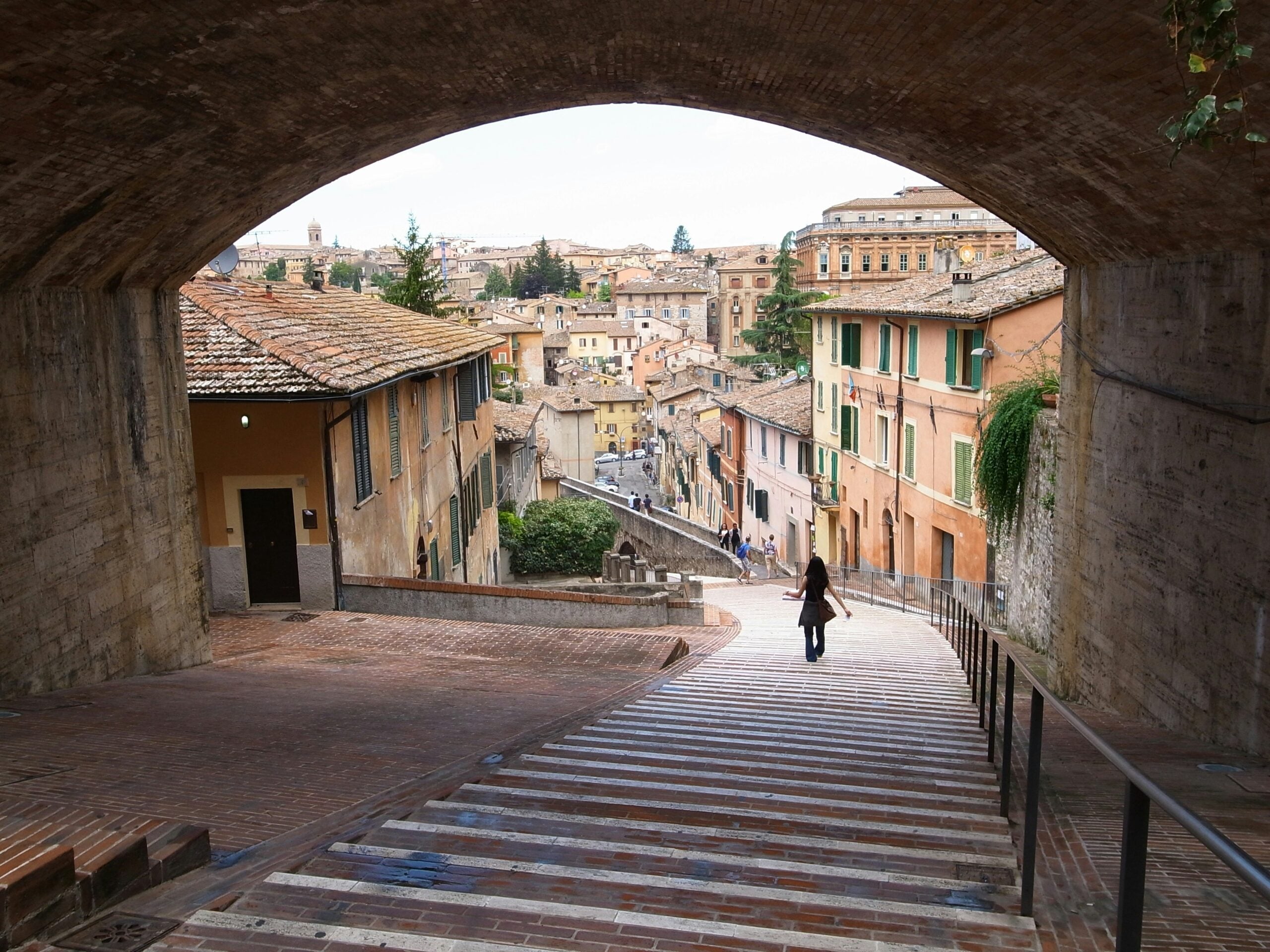 A woman walking down steep steps somewhere in Perugia, Italy, surrounded by historic stone buildings and lush greenery. She descends carefully, the ancient cobblestone steps leading her through a picturesque alleyway adorned with vibrant flowers and rustic lanterns. The scene captures the essence of Perugia's medieval charm and architectural beauty, highlighting the city's timeless allure and enchanting atmosphere.