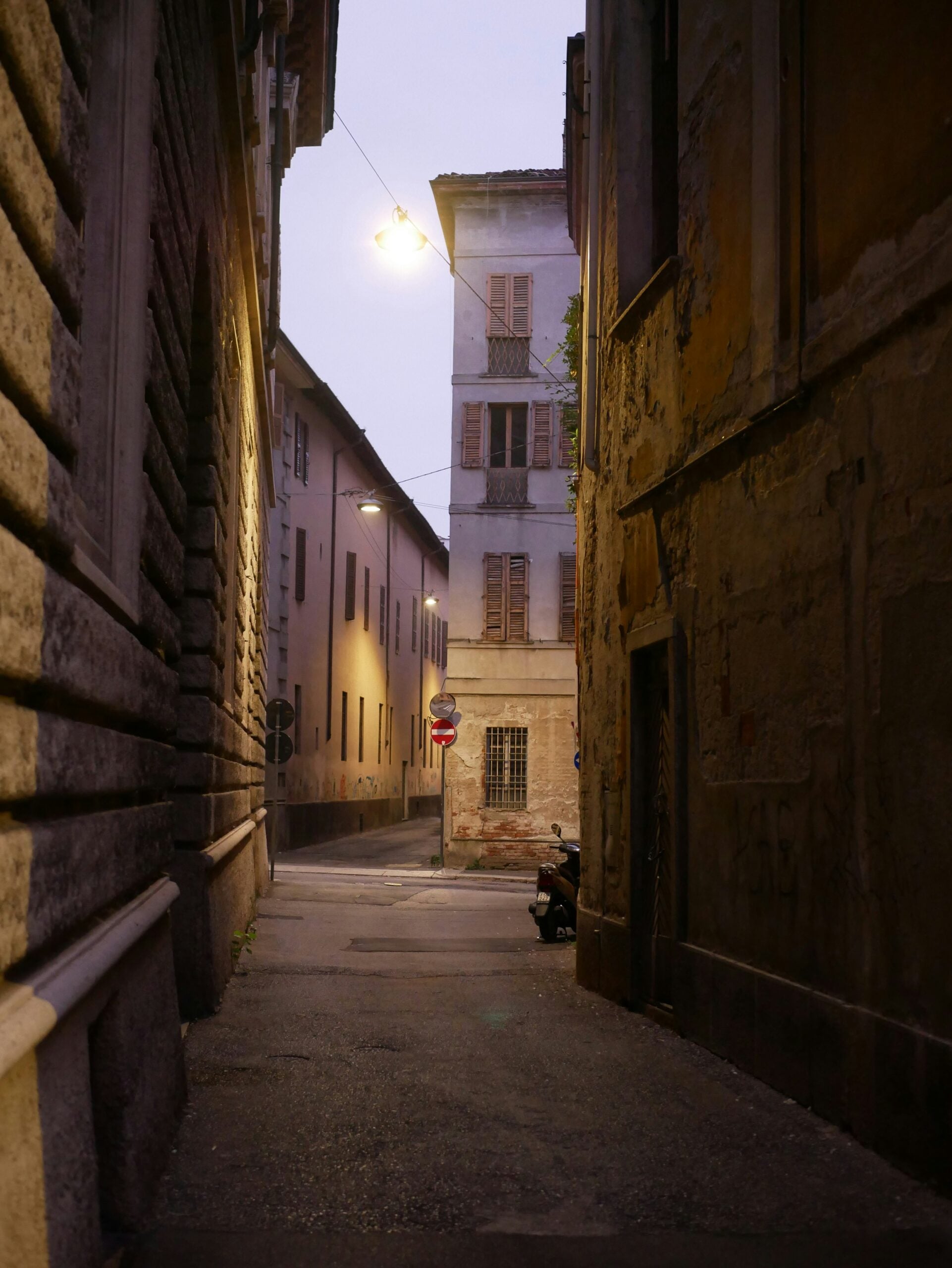 A serene photograph captures an empty street in the historic old city center of Cremona, Italy. The narrow cobblestone street is flanked by charming medieval buildings with colorful facades and ornate balconies. The quiet, early morning light casts gentle shadows, highlighting the intricate architectural details and wrought-iron lanterns. In the distance, the majestic Torrazzo tower of the Cremona Cathedral rises above the rooftops, adding a touch of grandeur to the tranquil scene. This peaceful moment in the heart of Cremona, Italy, invites exploration of its rich history and timeless beauty.