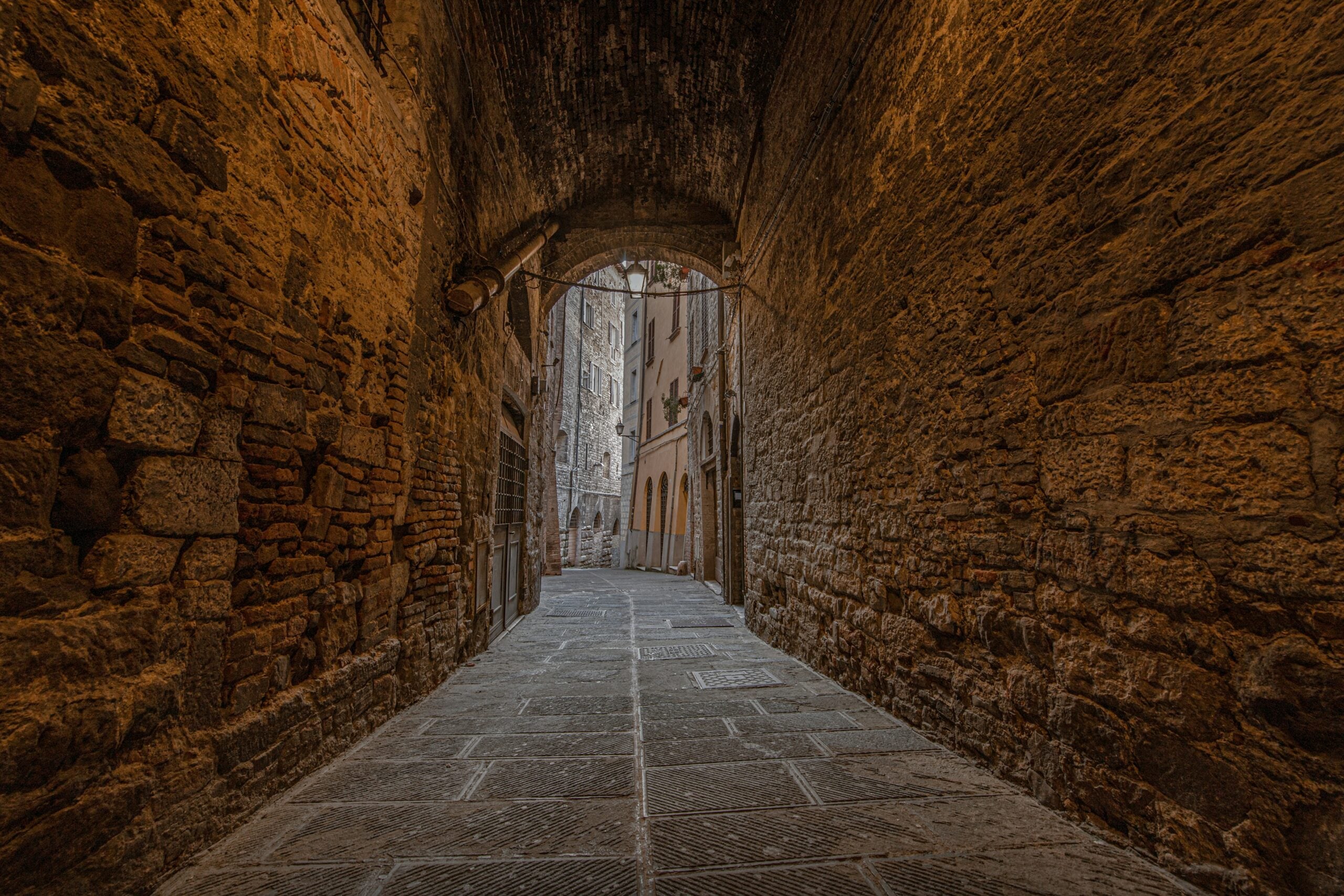 An old alley somewhere in Perugia, Italy, showcasing narrow, winding cobblestone paths flanked by ancient stone buildings with weathered facades. The alley is adorned with rustic lanterns, flowering plants in window boxes, and arched doorways, exuding a timeless charm. This scene captures the essence of Perugia's rich history and medieval architecture, providing a glimpse into the city's enchanting past.