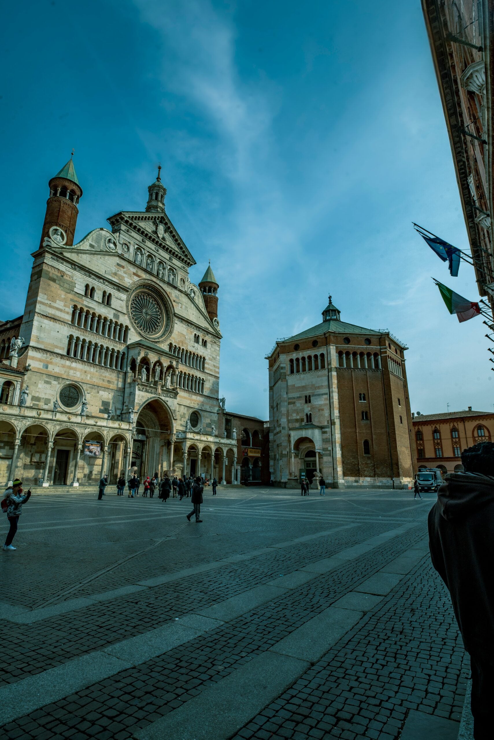 A stunning photograph captures the beautiful church and bustling piazza in Cremona, Italy. The elegant facade of the Cremona Cathedral, with its intricate Gothic and Romanesque design, stands majestically in the background. In the foreground, the lively piazza is filled with people enjoying outdoor cafes, historical statues, and charming cobblestone streets. The vibrant atmosphere of the square, coupled with the architectural beauty of the church, showcases the rich cultural heritage and inviting charm of Cremona, Italy, making it a captivating destination for travelers.