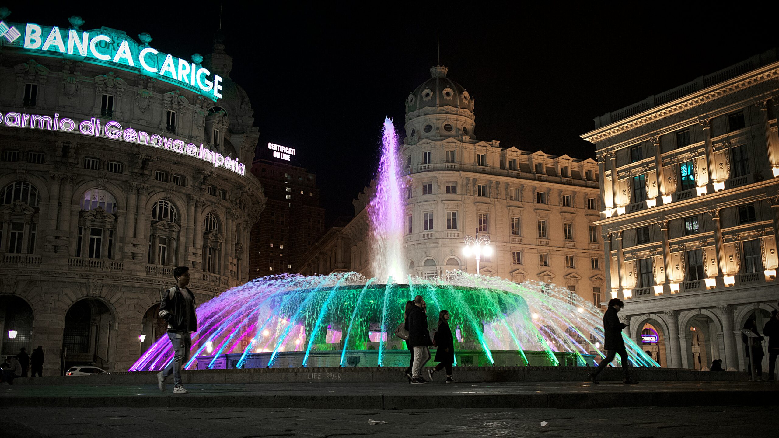 The photograph captures the main fountain in Piazza De Ferrari, Genoa, at night, beautifully illuminated against the dark sky. The fountain's graceful streams of water are highlighted by vibrant lights, casting a mesmerizing glow that enhances the grandeur of the scene. Surrounding the fountain, the historic buildings of the piazza are also lit up, their elegant facades reflecting the rich architectural heritage of the city. The soft, ambient lighting creates a serene and magical atmosphere, with the fountain as the centerpiece. This stunning night-time view of Piazza De Ferrari perfectly encapsulates the lively yet enchanting essence of Genoa, making it a must-see destination.