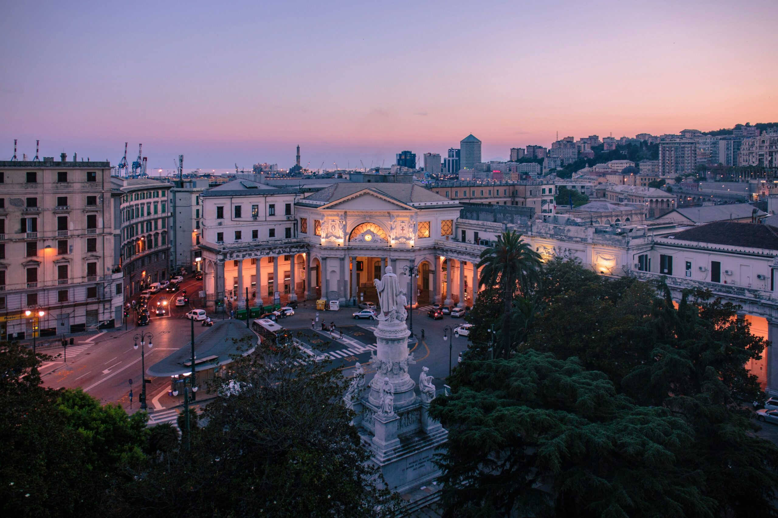 The photograph captures Genoa's main piazza, Piazza De Ferrari, at night, bathed in a warm, golden glow from the surrounding streetlights and illuminated buildings. The majestic central fountain, with its graceful arcs of water, takes center stage, reflecting the light and adding a sense of elegance to the scene. The historic architecture of the surrounding buildings, with their intricate facades and grandiose designs, creates a stunning backdrop against the deep blue night sky. The piazza is bustling with life, yet there is a serene ambiance that envelops the area, highlighting the timeless charm and vibrant nightlife of Genoa. This night-time view of Piazza De Ferrari perfectly encapsulates the lively yet enchanting atmosphere of the city, making it a must-see for visitors.