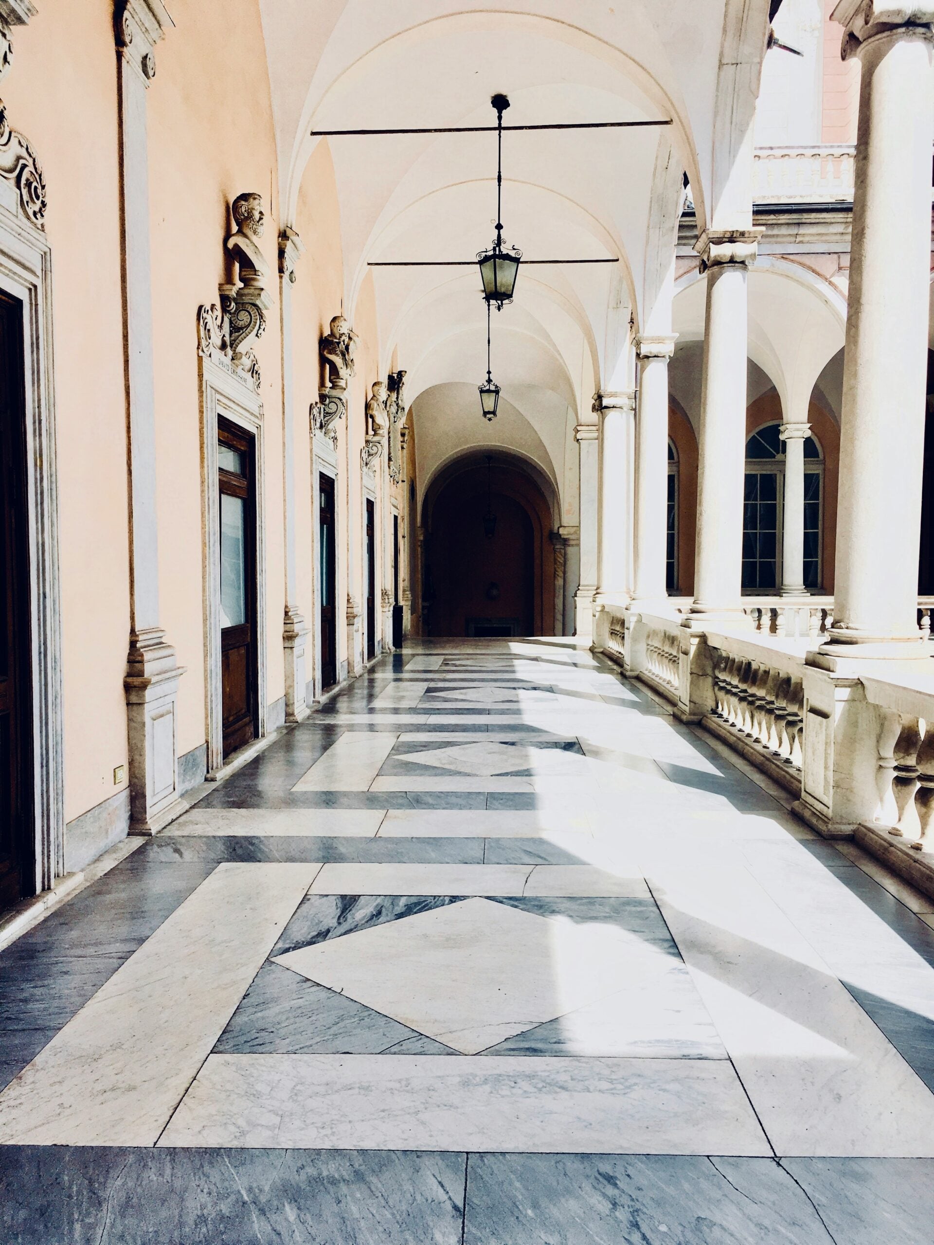 The photograph captures the interior of a beautiful marble walkway inside a grand building, likely in Genoa, Italy. The walkway is elegantly lined with polished marble columns and intricately designed arches that create a sense of grandeur and timeless elegance. The floor is adorned with intricately patterned marble tiles, reflecting the soft ambient light filtering in from large, arched windows or overhead lighting. The high ceiling, with its detailed moldings and ornate designs, adds to the majestic feel of the space. The walkway is quiet and serene, offering a peaceful passage through the heart of the building, showcasing the exquisite craftsmanship and architectural beauty typical of Genoa's historic structures.