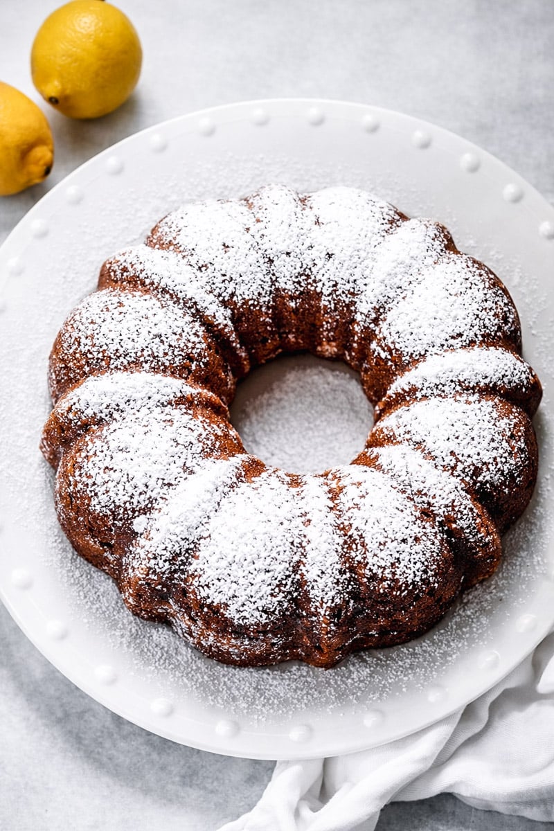 A round lemon ricotta cake dusted with powdered sugar sits on a white plate, with two whole lemons nearby on a light gray surface.