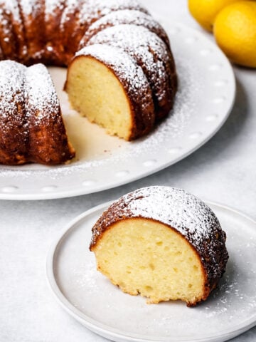 A slice of lemon ricotta cake dusted with powdered sugar sits on a white plate, with the rest of the cake and two lemons in the background.