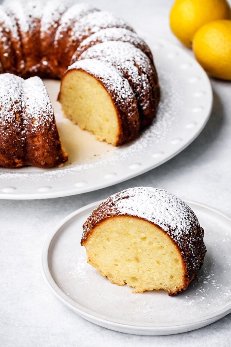 A slice of lemon ricotta cake dusted with powdered sugar sits on a white plate, with the rest of the cake and two lemons in the background.