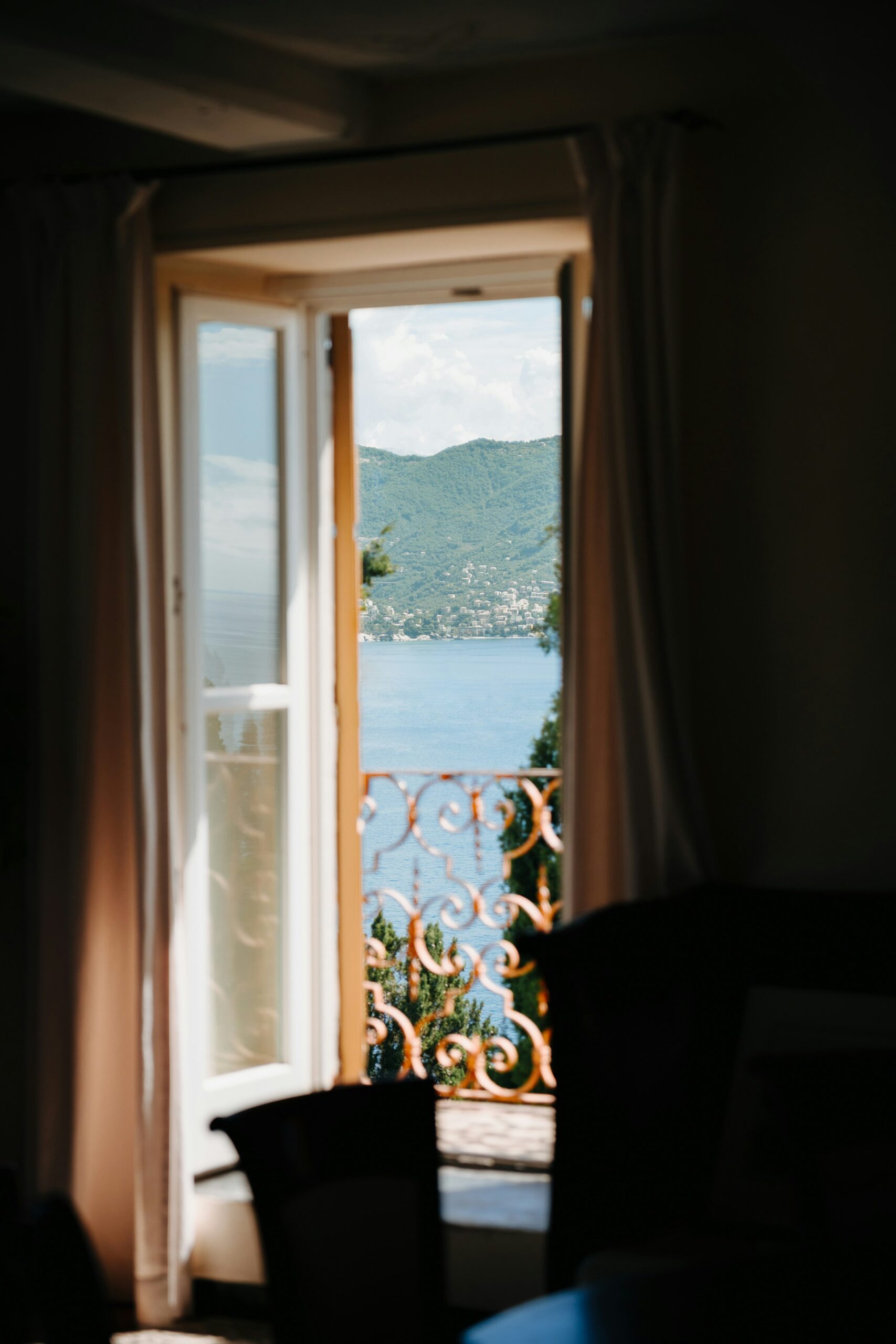 The photograph captures a stunning view looking out from a window at the bay in Portofino, Italy. Framed by the window's edges, the scene outside reveals the tranquil waters of the bay, where luxury yachts and colorful fishing boats are gently anchored. The window provides a perfect vantage point to admire the picturesque harbor, surrounded by vibrant, pastel-colored buildings that line the waterfront. Beyond the harbor, lush green hills rise, adding a natural, verdant backdrop to the idyllic scene. The sunlight dances on the water, creating sparkling reflections, and the overall view is serene and inviting, encapsulating the charm and elegance of Portofino from a cozy, intimate perspective.