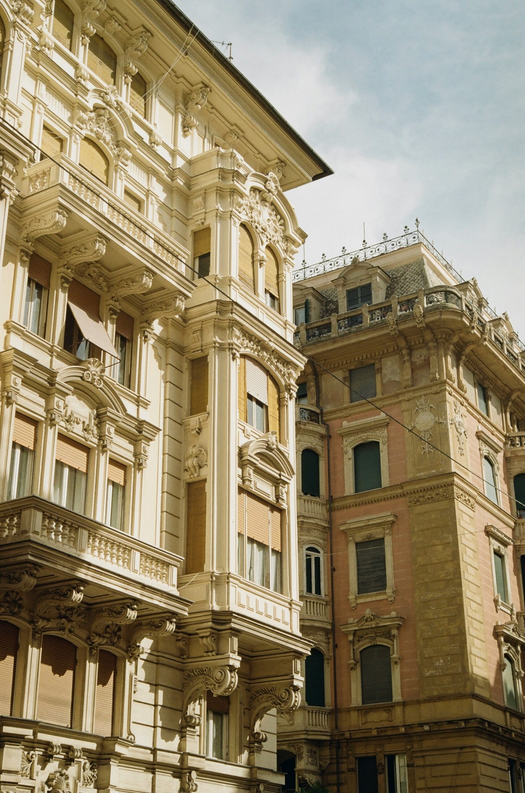 The photograph showcases the pretty architectural buildings of Genoa, Italy, highlighting the city's rich history and unique charm. The buildings are a mix of elegant palazzos and historic structures, featuring ornate facades adorned with intricate details such as decorative moldings, arched windows, and wrought-iron balconies. The colors of the buildings range from soft pastels to warm earth tones, typical of the Ligurian style. Some buildings are crowned with terracotta roofs, adding to the picturesque skyline. The scene is likely set on a quiet street or a bustling piazza, where the architectural beauty of Genoa