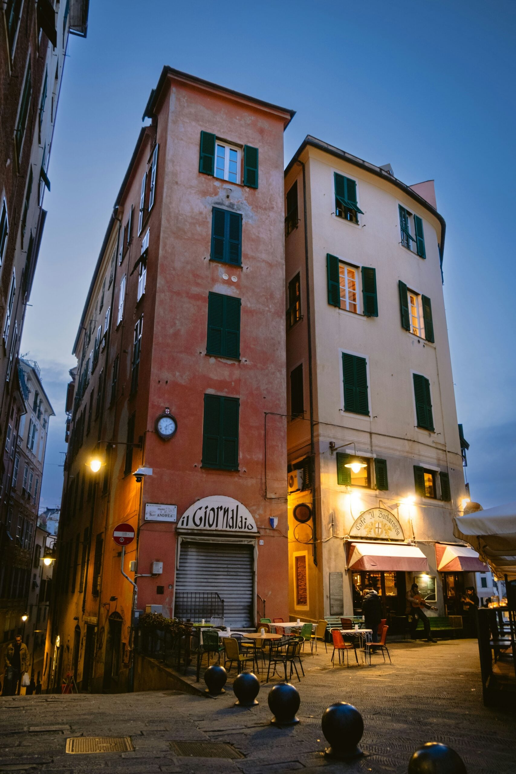 The photograph depicts residential buildings at night near the port of Genoa, Italy. The buildings, with their classic Italian architecture, stand tall against the night sky, their facades softly illuminated by streetlights. Warm, golden lights glow from the windows, creating a cozy and inviting atmosphere. Below, the port's waters reflect the shimmering lights, adding a sense of calm and serenity to the scene. The contrast between the bustling harbor and the tranquil residential area highlights the harmonious blend of urban life and maritime tradition in Genoa. This image captures the unique charm of living by the sea in this historic city.