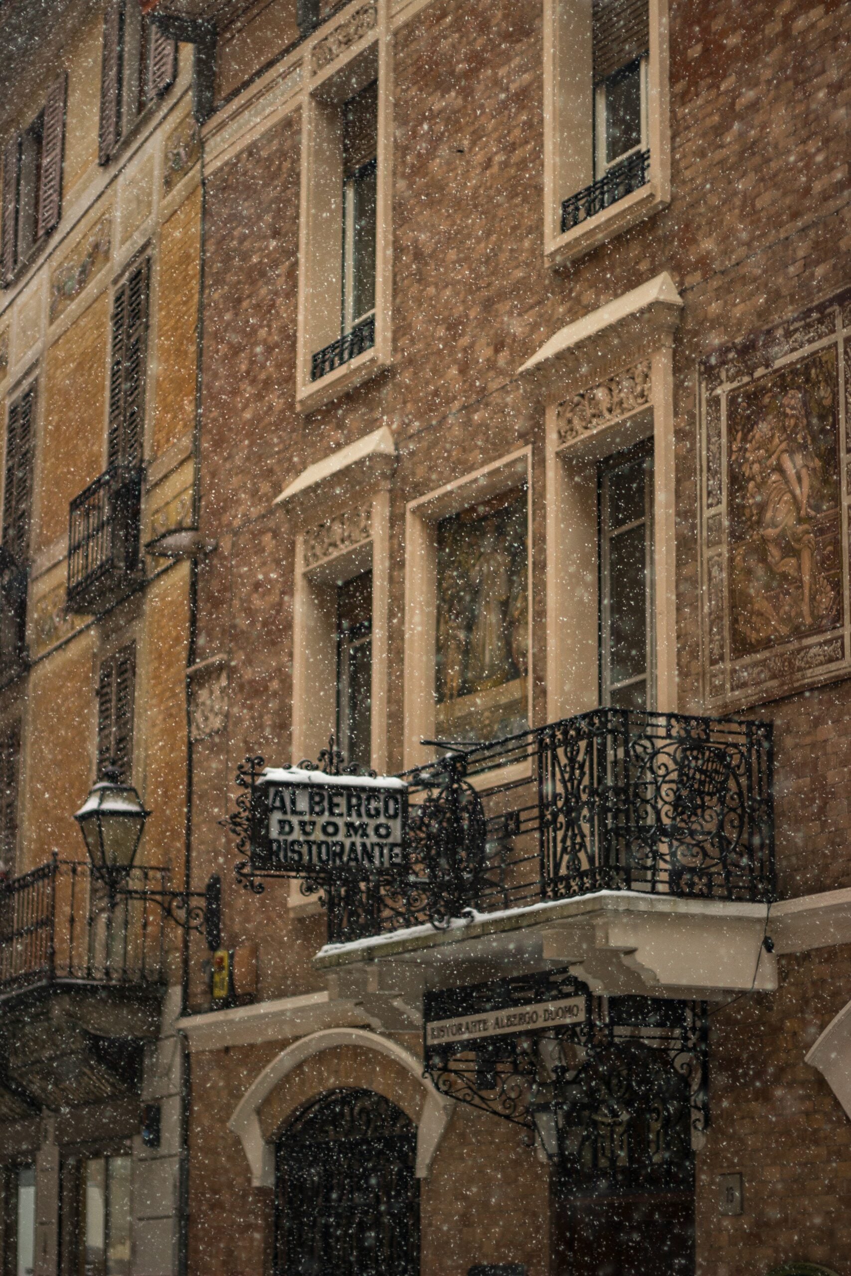 A serene photograph captures a picturesque snowfall over historic buildings in Cremona, Italy. The scene is transformed into a winter wonderland as a light layer of snow blankets the rooftops, streets, and the intricate facades of the medieval structures. The soft white snow contrasts beautifully with the warm tones of the ancient brick and stone buildings. Delicate snowflakes continue to fall, adding a magical touch to the quiet atmosphere. The iconic Torrazzo tower rises majestically in the background, its details softened by the snowfall, creating a charming and tranquil winter scene in the heart of Cremona, Italy.