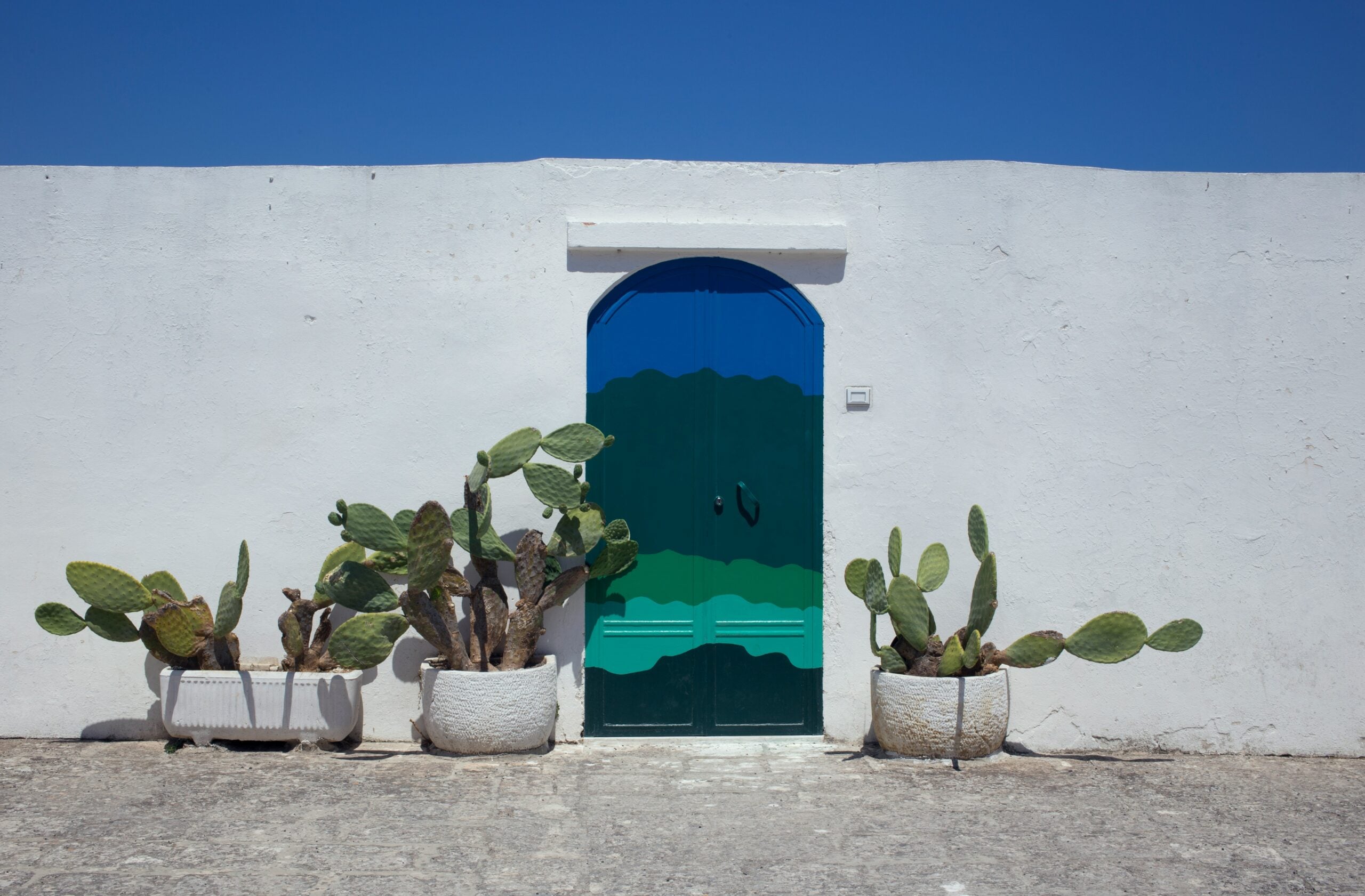 The photograph showcases the famous blue door in Ostuni, Italy, set against the town's characteristic whitewashed walls. This iconic door, painted a vivid azure, stands out as a vibrant focal point amidst the stark white backdrop. Surrounding the door, delicate stonework and rustic details add to the picturesque and charming aesthetic. This famous blue door is a beloved symbol of Ostuni, often referred to as the "White City," capturing the unique blend of color and architectural beauty that defines this enchanting town in Puglia. The image highlights the timeless allure and distinctive character of Ostuni, Italy.