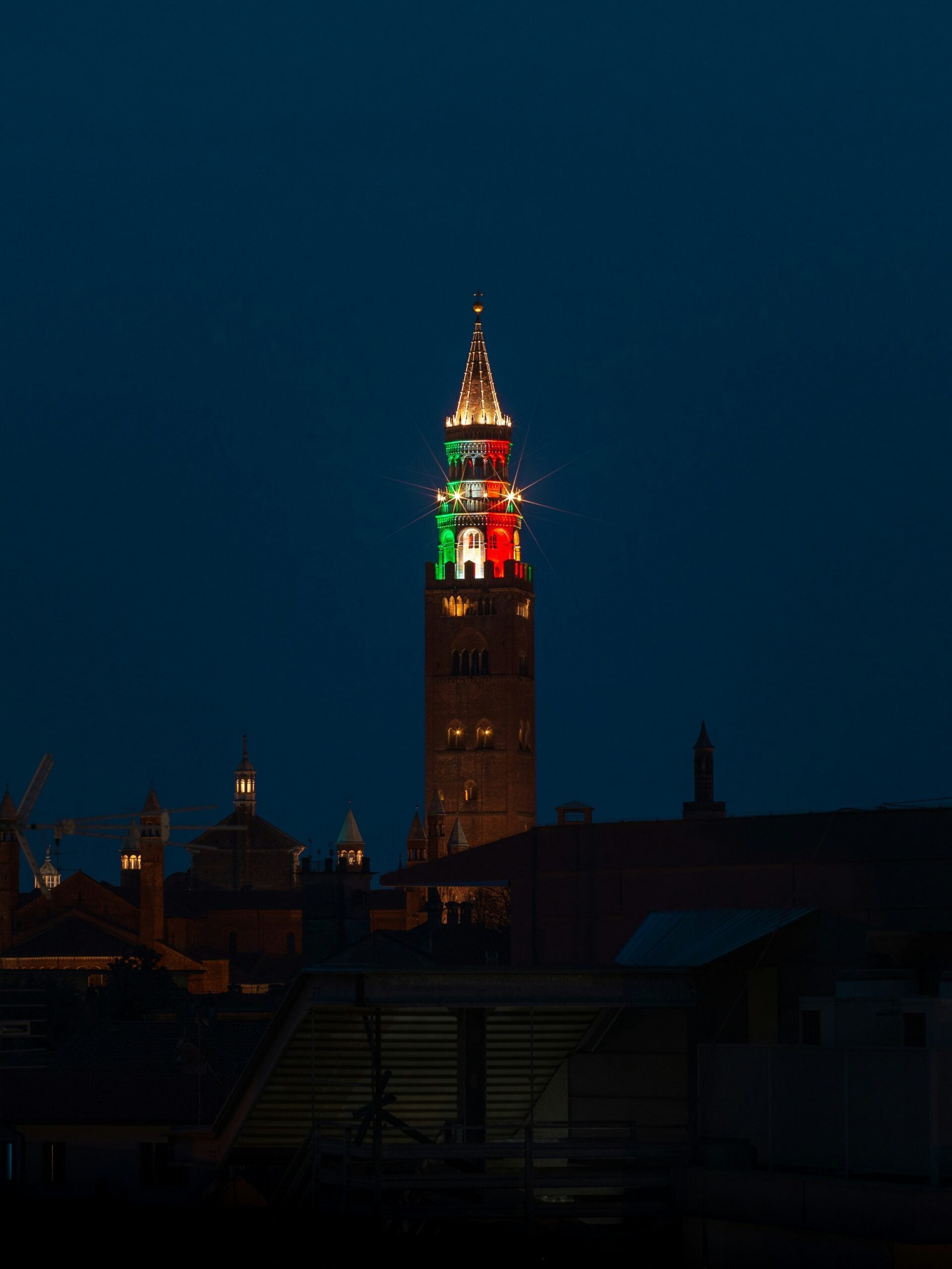 A captivating nighttime photograph showcases the large ancient Torrazzo tower in Cremona, Italy, brilliantly illuminated with the colors of the Italian flag. The tower's impressive height and historic brickwork are bathed in vibrant green, white, and red lights, creating a striking contrast against the dark night sky. The Italian flag colors cast a patriotic glow, highlighting the tower's architectural details and historical significance. Below, the softly lit square and surrounding buildings add to the enchanting atmosphere, making this night view of the Torrazzo in Cremona, Italy, a spectacular and memorable sight.
