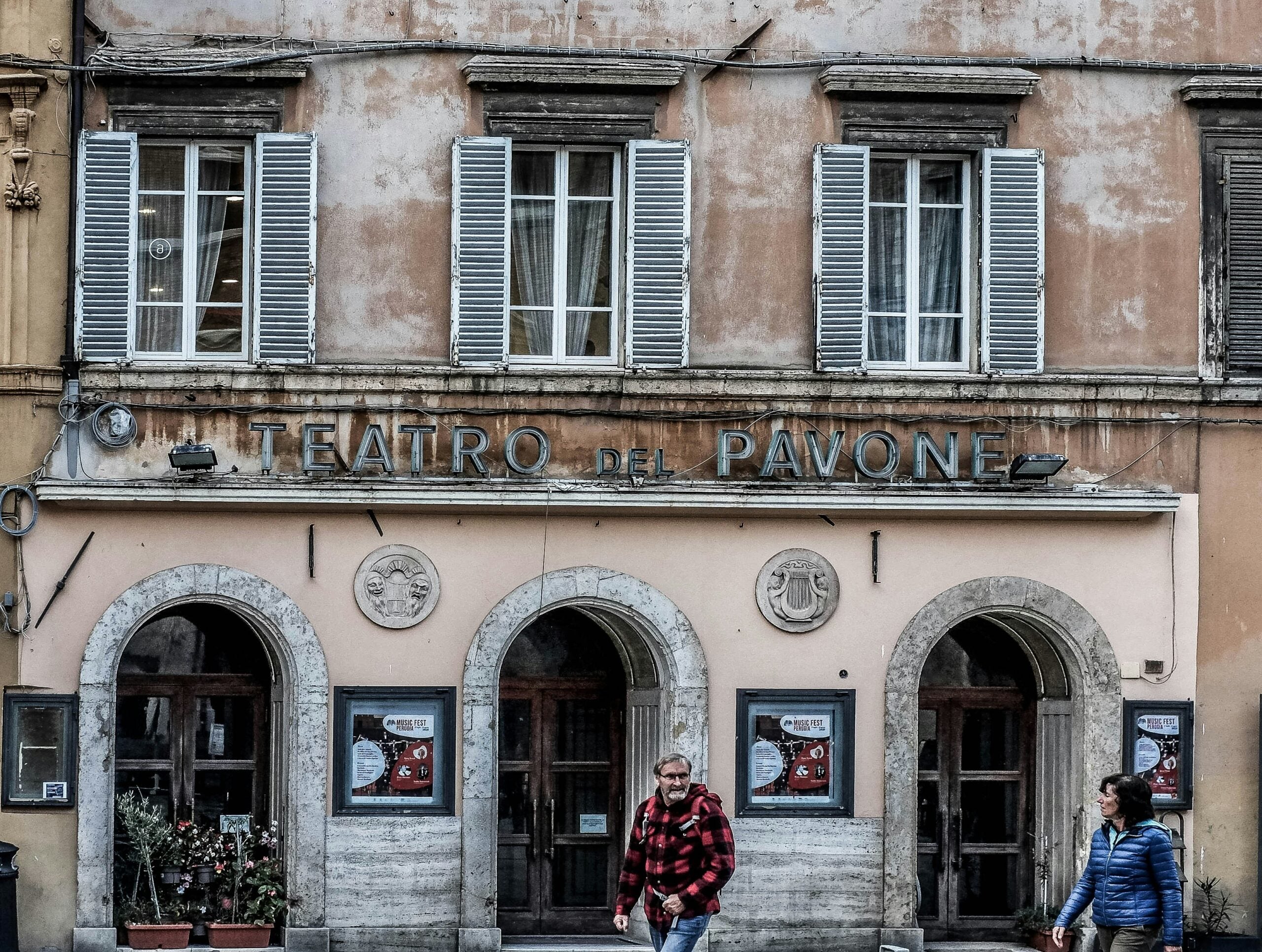 Tourists gather outside an old theater in Perugia, Italy, admiring its grand facade with ornate stone carvings and historic architectural details. The building's arched entrance and large wooden doors add to its majestic appearance. Visitors take photos and read informational plaques, immersing themselves in the rich cultural heritage of Perugia. The scene captures the vibrant atmosphere and timeless charm of this historic Italian city.