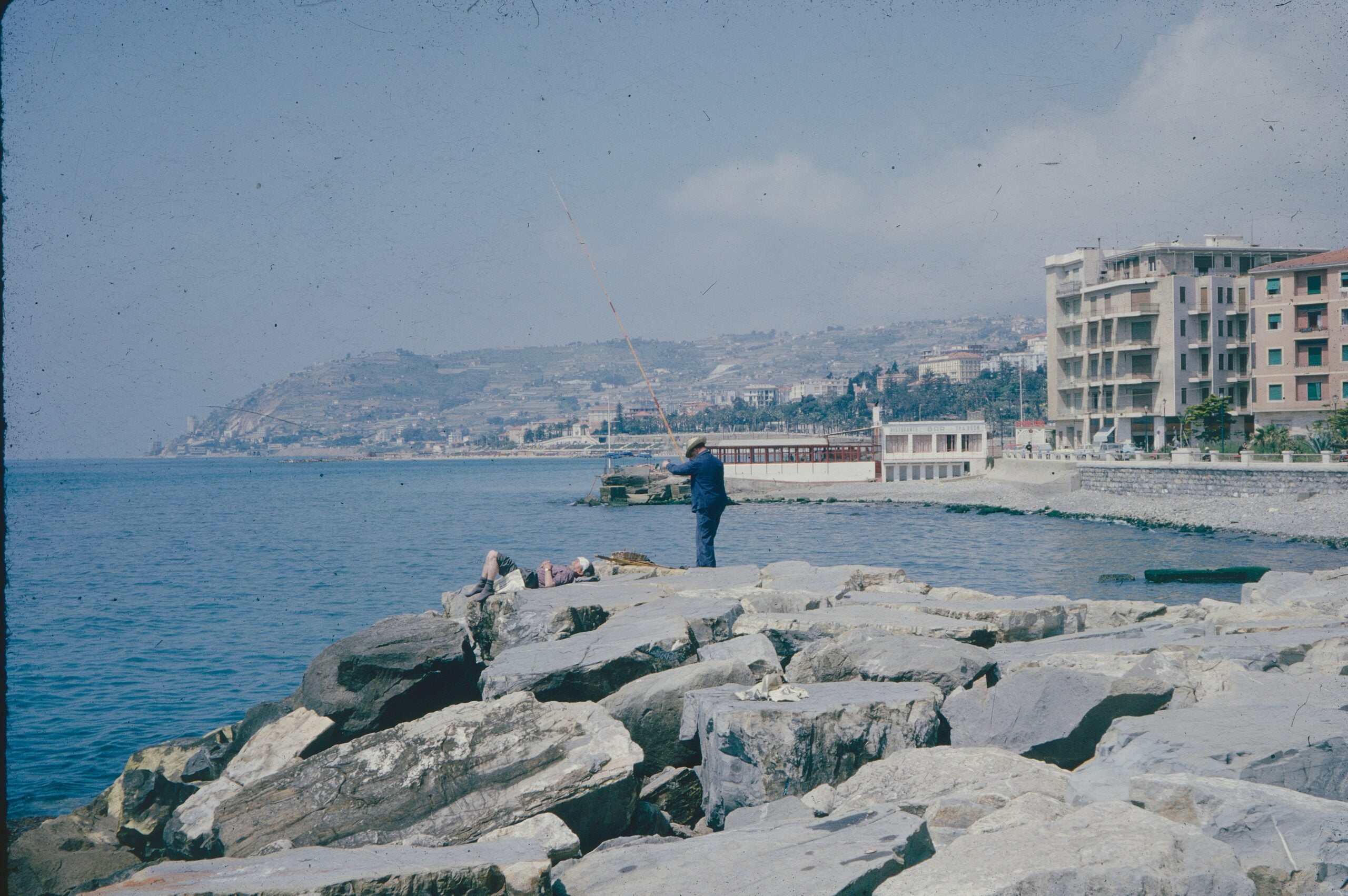 A faded color photograph from 1967 depicting a man fishing in San Remo, Italy. The image captures a moment of quiet reflection, with the man standing on a rocky shoreline, casting his line into the calm Mediterranean waters. His attire, typical of the era, adds a nostalgic touch to the scene. The colors of the photograph have softened over time, giving it a vintage, timeless feel. The backdrop features the serene sea and hints of the coastal landscape, encapsulating a peaceful day in San Remo, Italy, during the late 1960s.