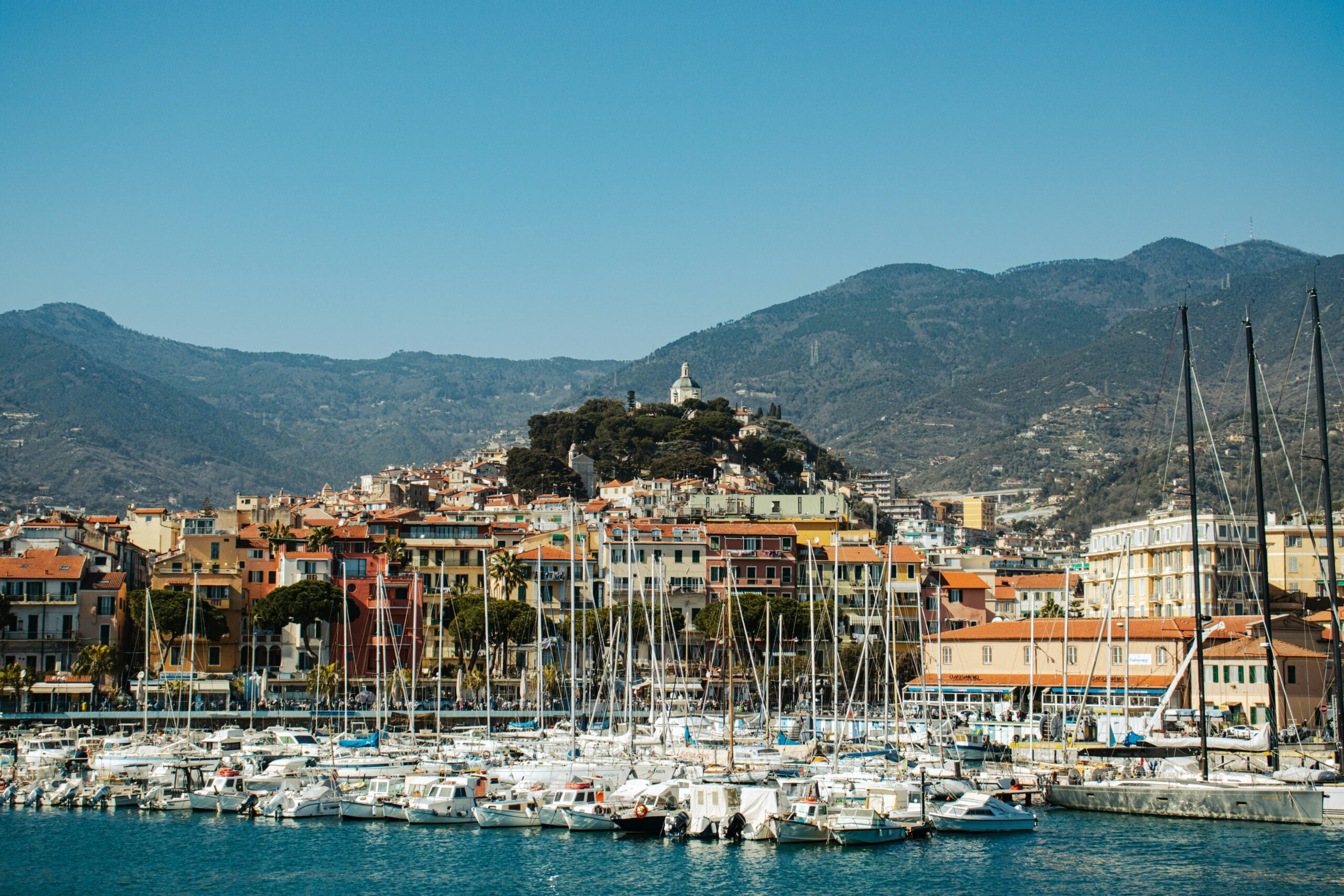 A picturesque view of San Remo in the Province of Imperia, Italy, featuring an array of sailboats docked in the harbor on a bright, sunny day. The sailboats, with their masts stretching towards the clear blue sky, create a vibrant scene that highlights the coastal charm of this beautiful Italian town. The calm waters of the harbor reflect the boats, adding to the serene atmosphere. This image captures the essence of San Remo, Italy, a popular destination known for its maritime beauty and Mediterranean allure.