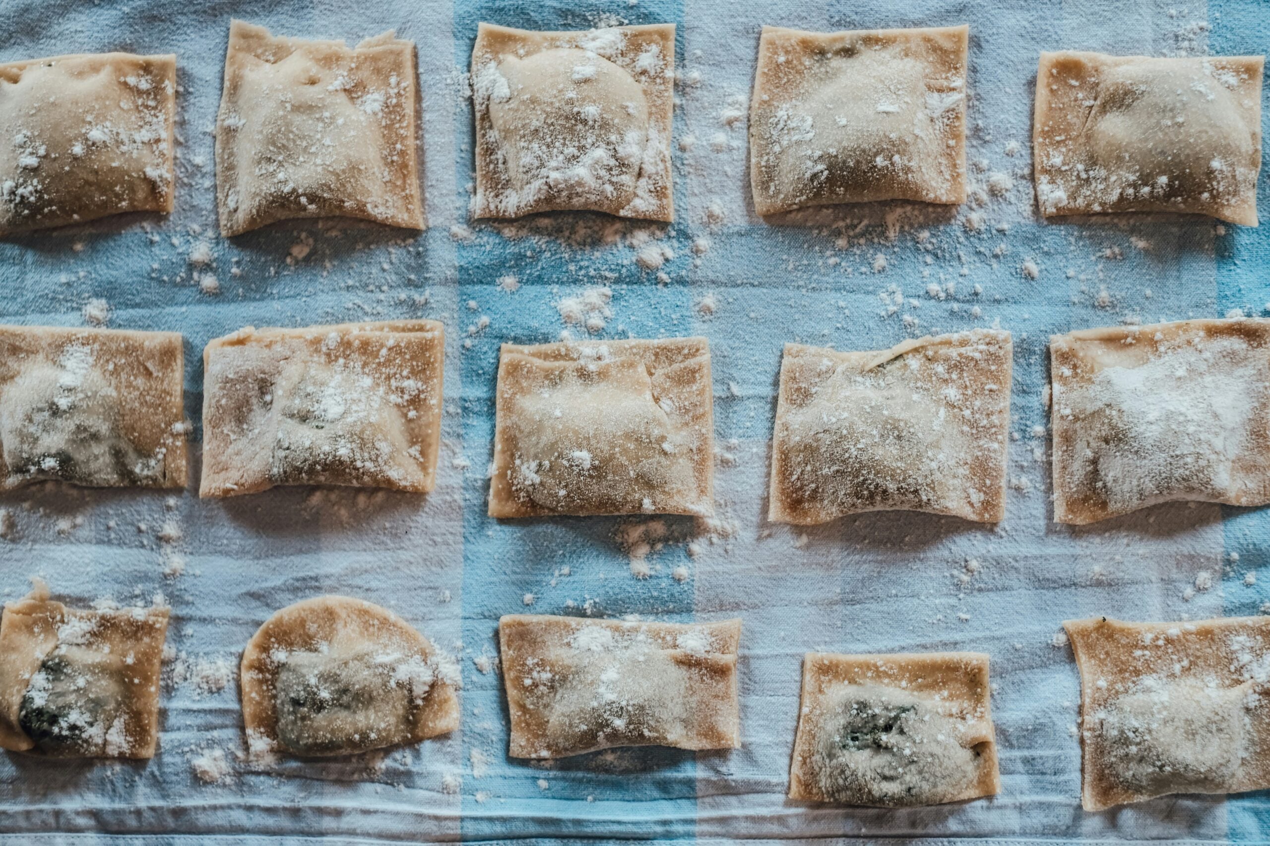 A photograph captures the rustic charm of handmade Tortelli pasta from Reggio Emilia, Italy, drying on a wooden table. The golden pasta squares are carefully arranged, their edges crimped with precision, showcasing the artistry involved in traditional Italian cooking. The image highlights the fresh, doughy texture of the pasta, with hints of flour dusted on the table surface. This scene reflects the authentic culinary heritage of Reggio Emilia, Italy, where making Tortelli is a time-honored tradition passed down through generations, celebrated for its simplicity and rich flavors.