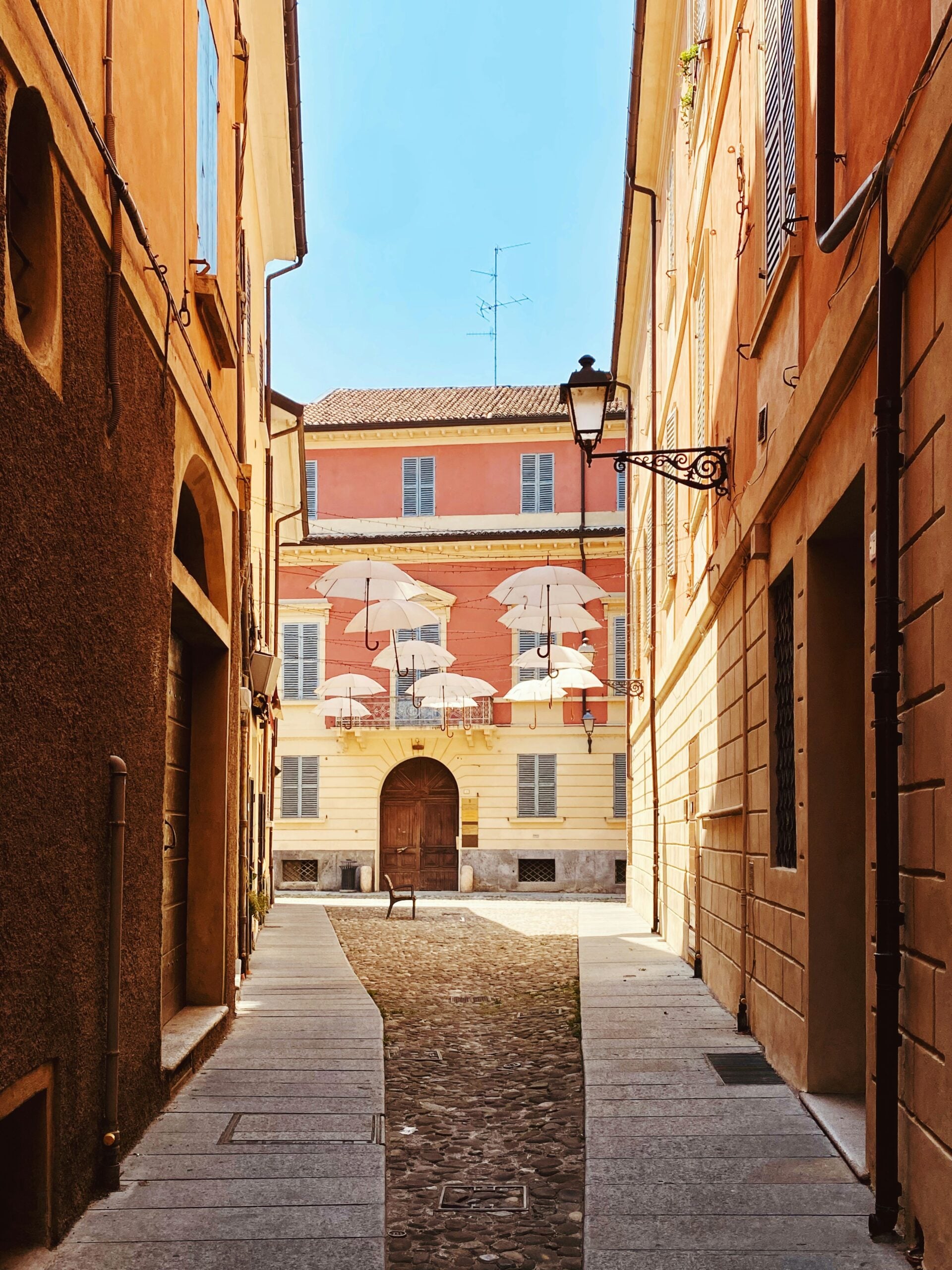 A photograph of Via Palazzolo 2B–5A in Reggio Emilia, Italy, captures a charming street scene in this historic city. The image showcases the classic Italian architecture of the buildings, with their pastel-colored facades, shuttered windows, and wrought-iron balconies. The cobblestone street adds to the old-world charm, while potted plants and flowers bring a touch of color and life to the scene. This picturesque street reflects the everyday beauty and timeless appeal of Reggio Emilia, Italy, offering a glimpse into the local lifestyle and architectural heritage.