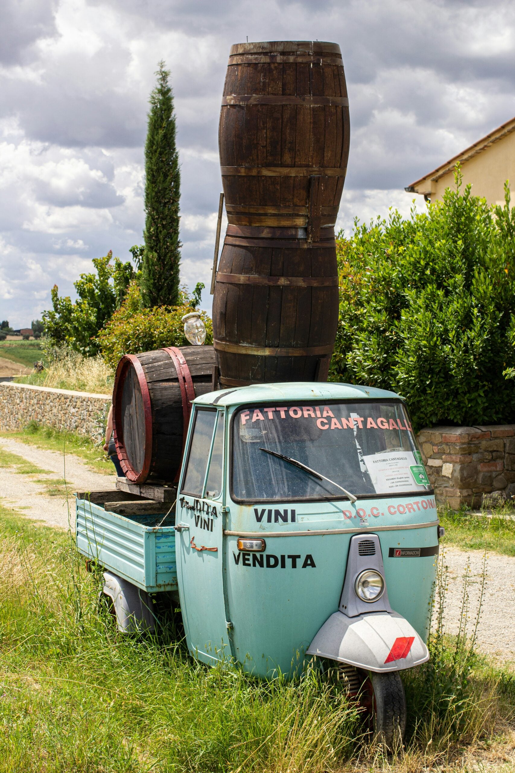 The image features a set of wine barrels carefully stacked atop a vintage blue Piaggio Ape, a classic three-wheeled vehicle, at Fattoria Cantallo in Arezzo, Italy. The Piaggio Ape, with its rustic charm and iconic design, contrasts beautifully with the rich wooden tones of the wine barrels. The scene is set against the backdrop of the picturesque Tuscan countryside or a traditional vineyard setting, highlighting the blend of old-world craftsmanship and local heritage. This image captures the essence of Italian culture, combining the love of wine with the charm of vintage vehicles, set in the idyllic surroundings of Arezzo, Italy.