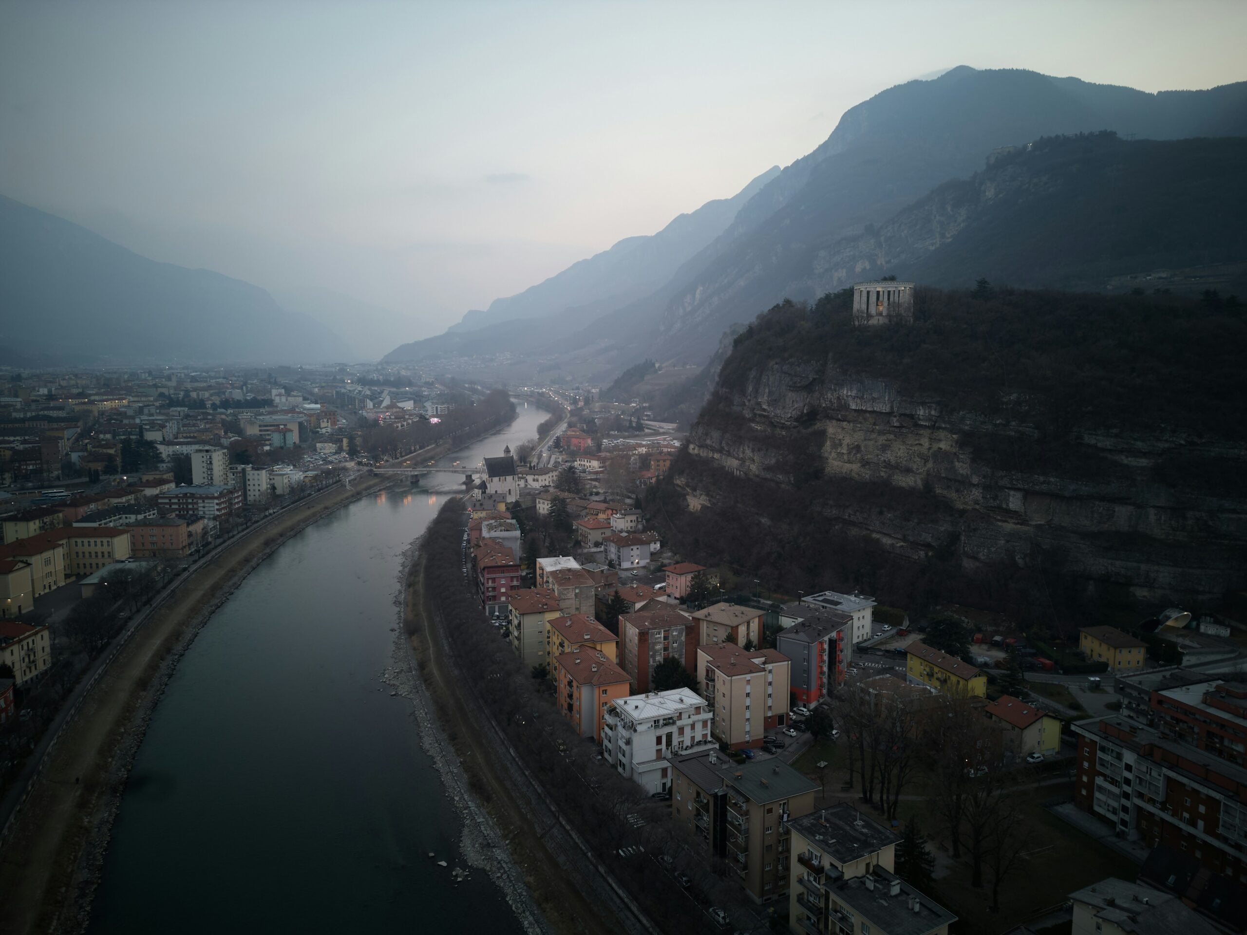 An aerial view of Trento, Italy, on a cloudy day, showcasing the city's intricate layout nestled in a valley surrounded by rolling hills. The Adige River winds gracefully through the heart of Trento, its waters reflecting the muted light filtering through the overcast sky. The city's blend of historic buildings, bridges, and green spaces is visible from above, with the surrounding mountains partially shrouded in mist, adding a touch of mystery to the scene. This aerial perspective highlights the unique geography and the seamless integration of urban life with the natural landscape in Trento, Italy.