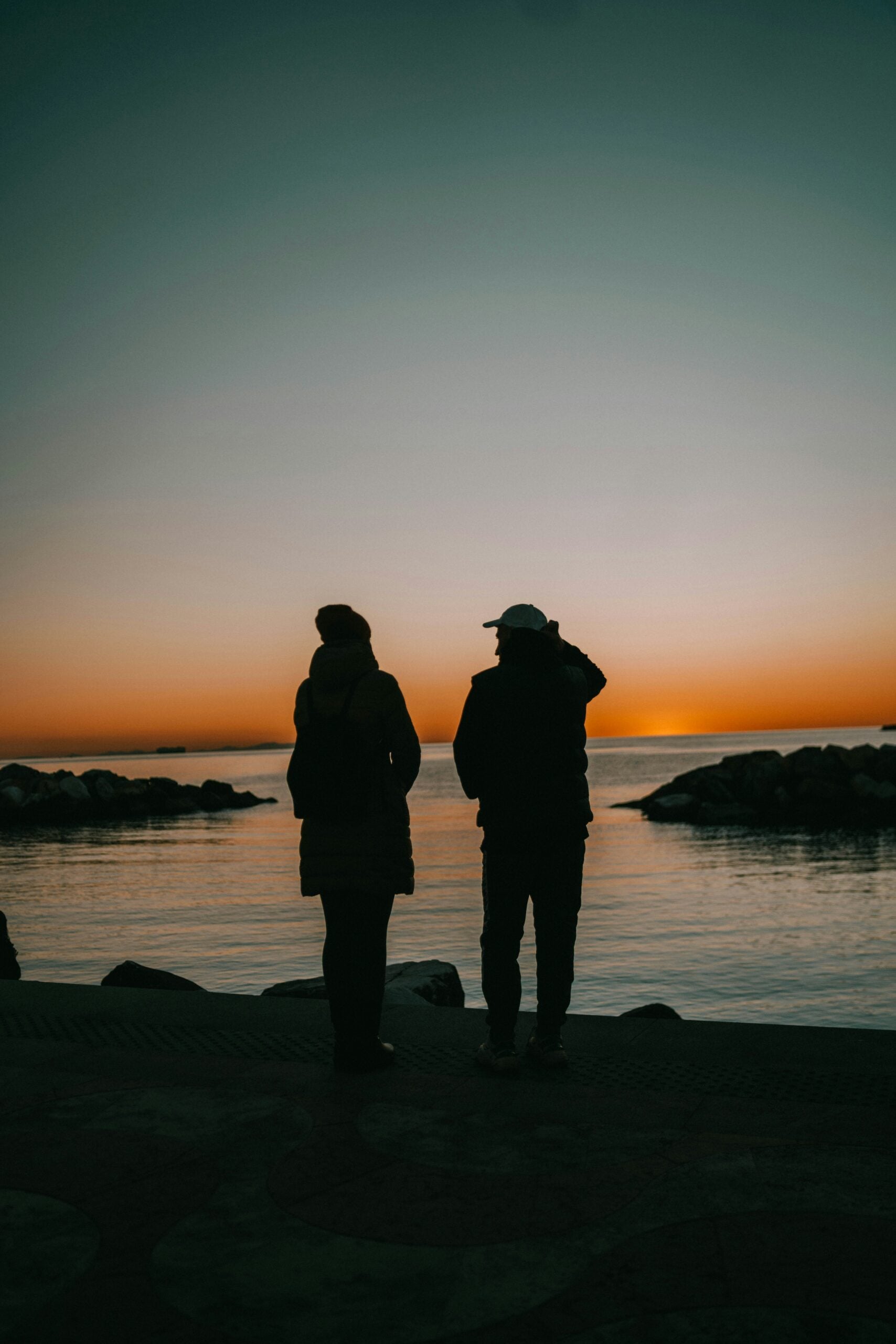 A couple stands side by side, gazing at the breathtaking sunset. The sky is painted with warm hues of orange, pink, and purple as the sun dips below the horizon. The serene moment captures the couple's silhouettes softly illuminated by the fading light. The tranquil waters in the foreground reflect the vibrant colors of the sky, adding to the peaceful ambiance. This image beautifully conveys a sense of romance and tranquility, as the couple shares this quiet, intimate moment with the natural beauty of the sunset as their backdrop.