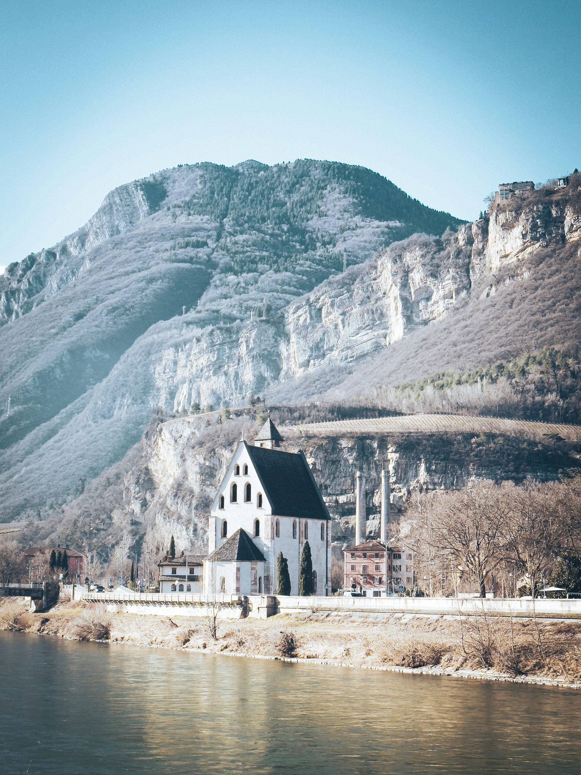 A serene winter scene featuring a small, picturesque church by the edge of a tranquil lake in Trento, Italy. The church, with its simple yet charming architecture, is partially dusted with snow, adding to the peaceful ambiance. The lake's surface reflects the soft light of a cloudy winter day, with the surrounding landscape blanketed in snow, creating a quiet, almost ethereal atmosphere. Tall, snow-covered trees frame the church, and the distant mountains rise softly in the background, completing the idyllic winter setting. This peaceful scene captures the tranquil beauty of winter in Trento, Italy.