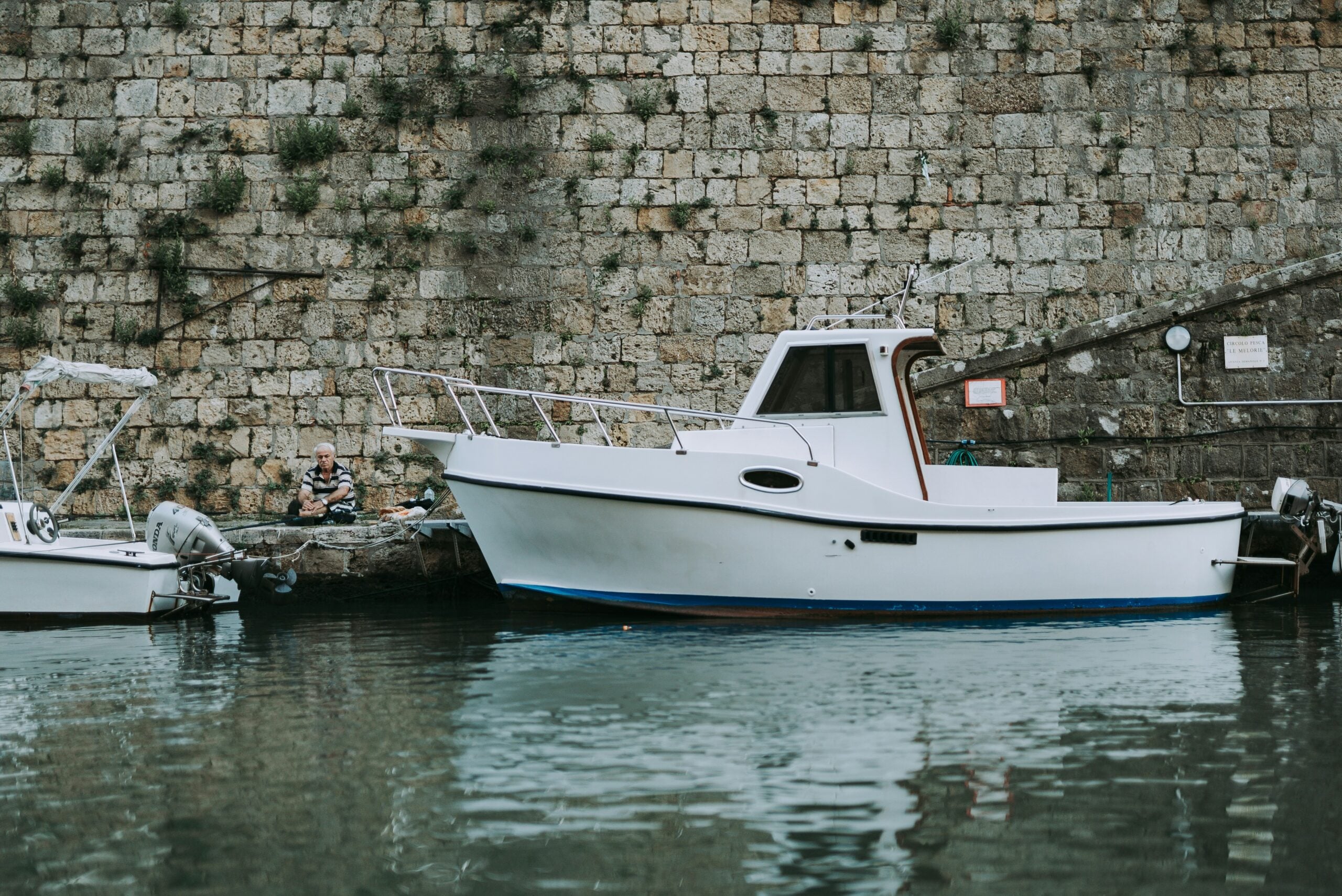 A tiny boat is gently moored along the edge of a narrow canal, creating a peaceful and picturesque scene. The boat, with its simple design and weathered paint, floats calmly on the still water, which reflects the surrounding buildings and soft light of the sky above. The canal is lined with cobblestone paths and historic buildings, their facades adding to the charm of the setting. The quiet atmosphere suggests a moment of tranquility in an otherwise bustling area, highlighting the serene beauty of this hidden corner in a coastal town, possibly Livorno, Italy.