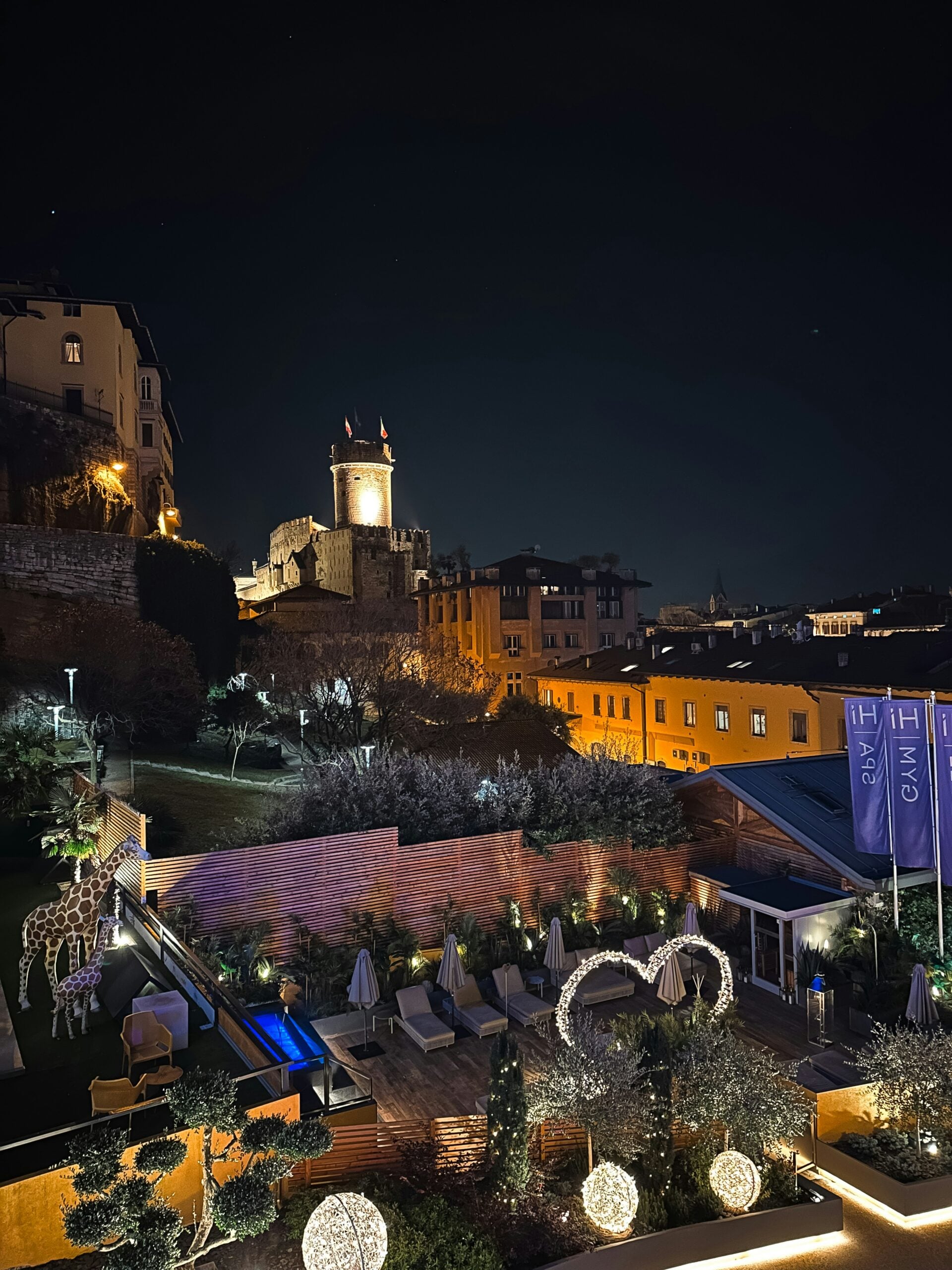 A captivating nighttime view from a hotel room overlooking the hilly section of Trento, Italy. The scene is bathed in the soft glow of streetlights and the warm lights from homes scattered across the hills. The undulating landscape creates a sense of depth, with the lights twinkling like stars against the dark silhouette of the hills. In the distance, the outlines of the mountains are barely visible under the night sky, adding a serene and peaceful ambiance. This enchanting view offers a tranquil and intimate glimpse of Trento, Italy, as the city rests under the quiet night.
