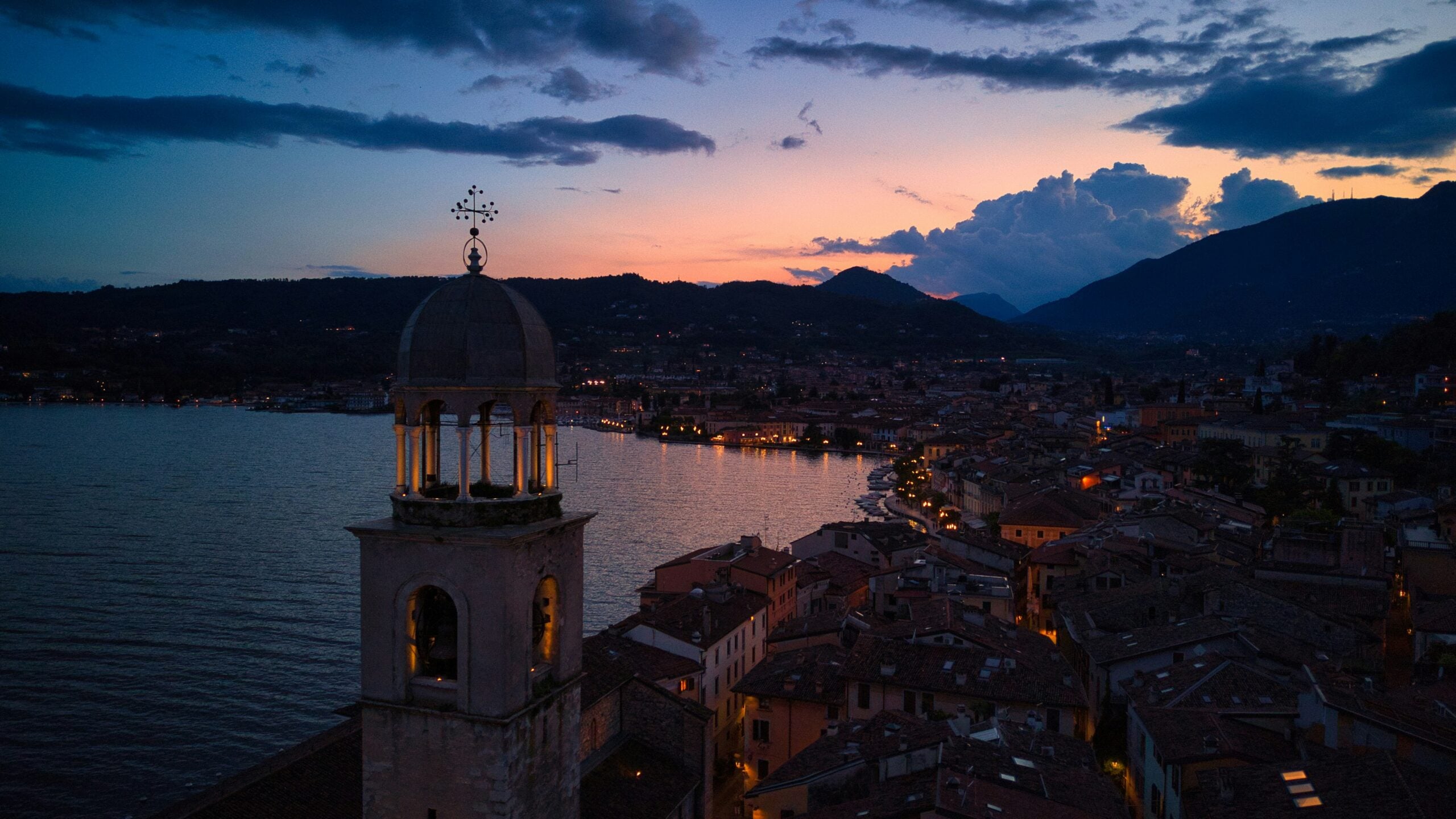 Aerial view of Brescia at sunset, capturing the early evening glow over water, highlighting the city's beautiful skyline against a backdrop of fading light. This photograph showcases the peaceful transition from day to night, with soft orange and pink hues reflecting off the water, offering a breathtaking perspective of this historic Italian city.
