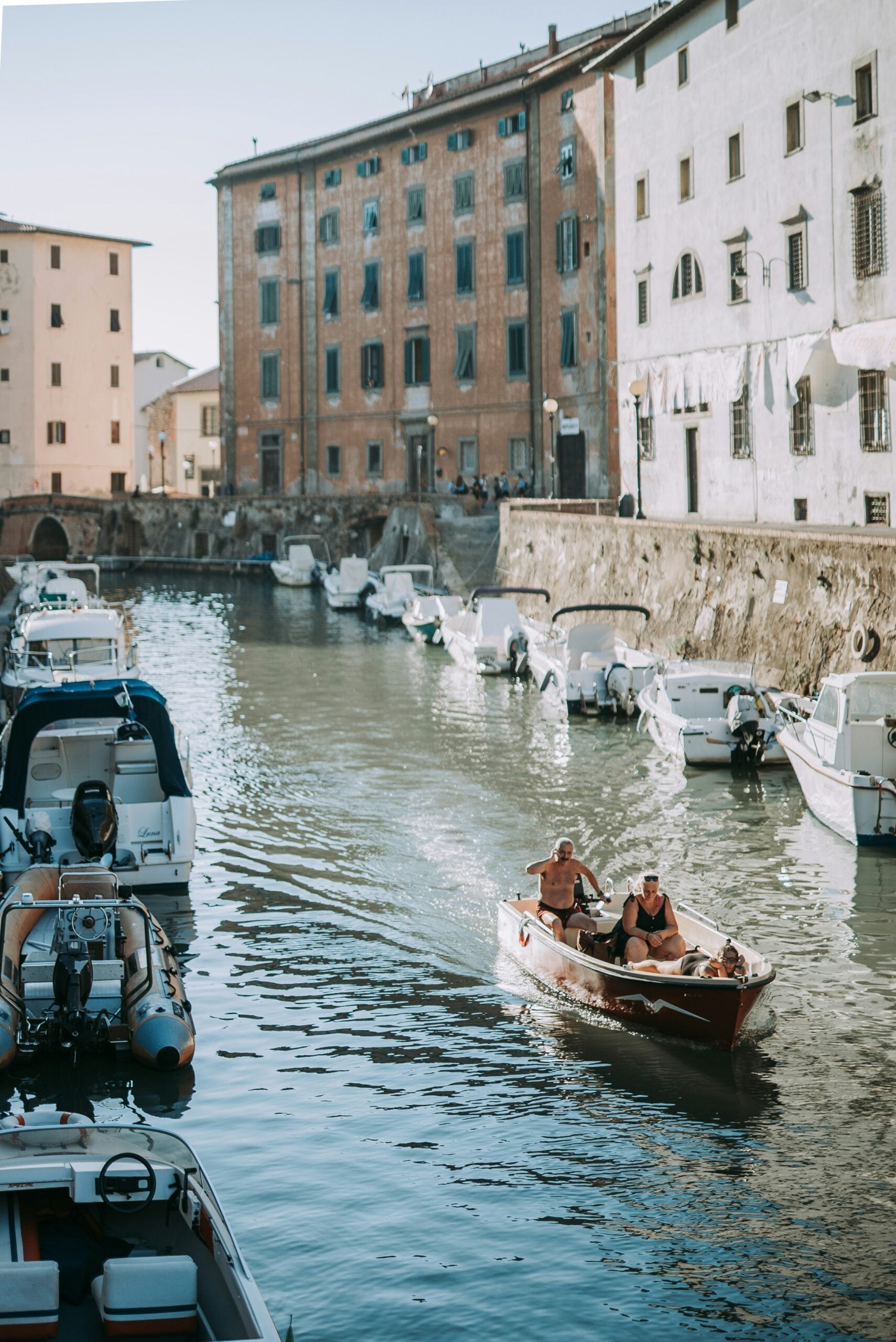 An elderly couple is seen taking a small boat out on a tranquil canal. The boat gently glides through the calm water, creating soft ripples as they move. The couple, both wearing light, comfortable clothing, sit close together, with the man carefully guiding the boat while the woman enjoys the peaceful surroundings. The canal is lined with charming old buildings, their reflections mirrored in the water, adding to the serene atmosphere. This image captures a tender moment of togetherness and adventure, highlighting the enduring bond between the couple as they enjoy a quiet journey on the canal.